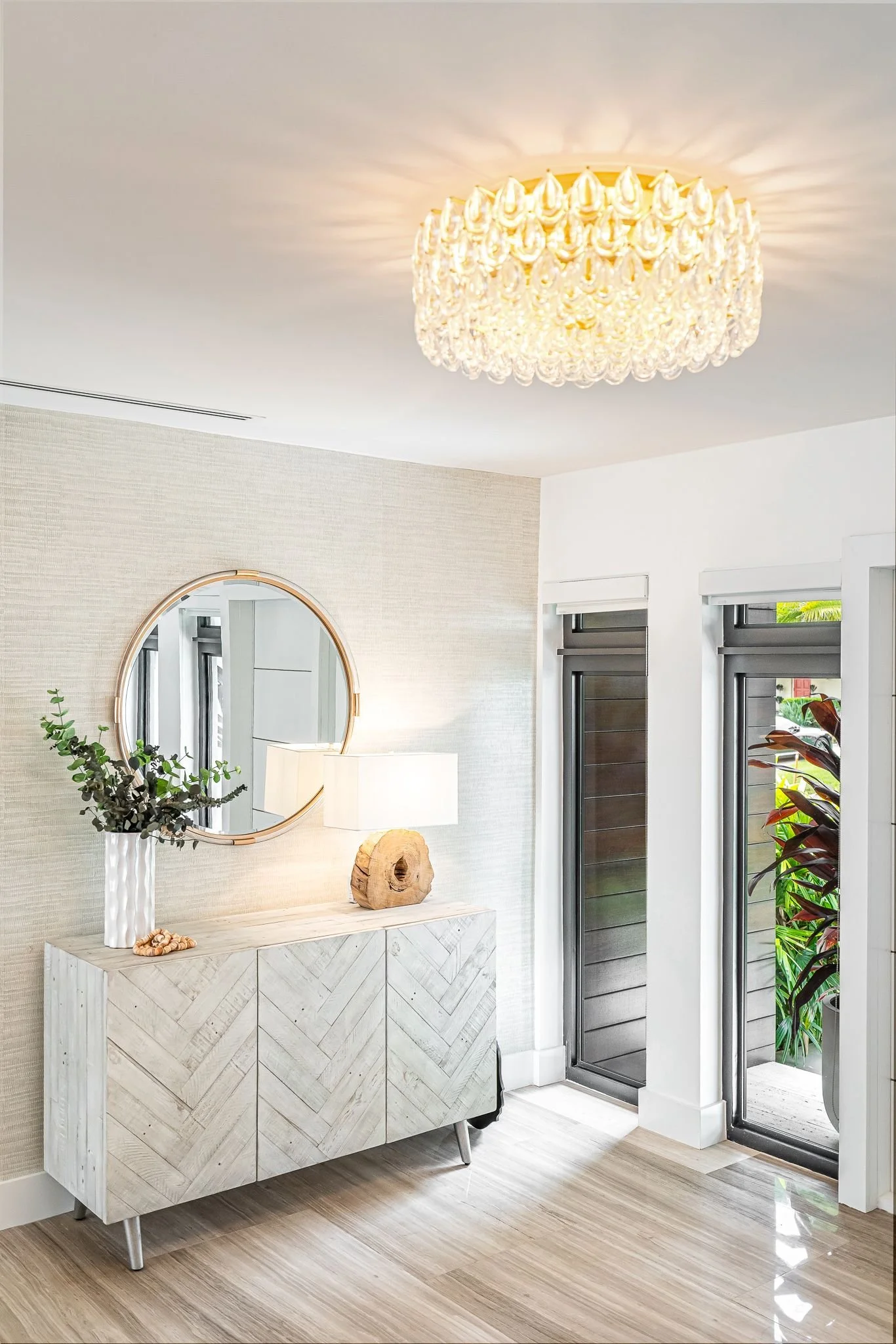 Modern entryway with a chandelier ceiling light, a round mirror, a sideboard with decorative items, and glass sliding doors leading outside.