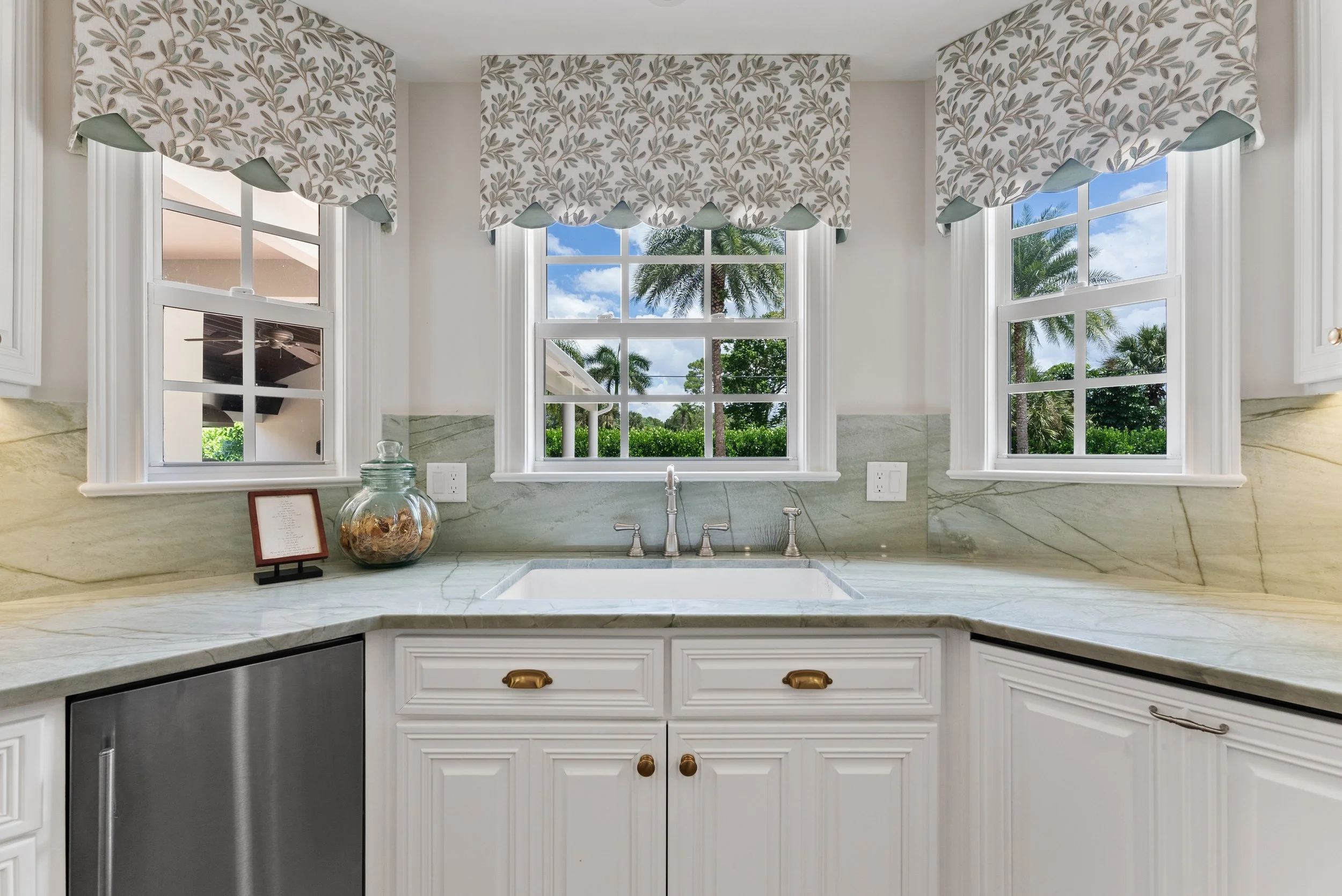 Kitchen sink area with three windows showing palm trees and blue sky, white cabinetry, marble countertop, and decorative window valances.