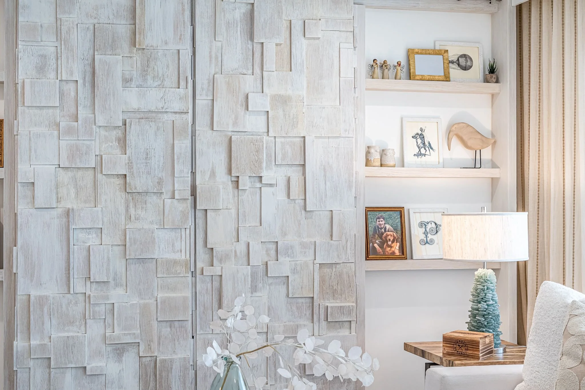 Interior living space with a textured white wall panel, open shelving displaying decorative items and photos, a table lamp with a blue textured base, and a beige armchair with a textured pillow.