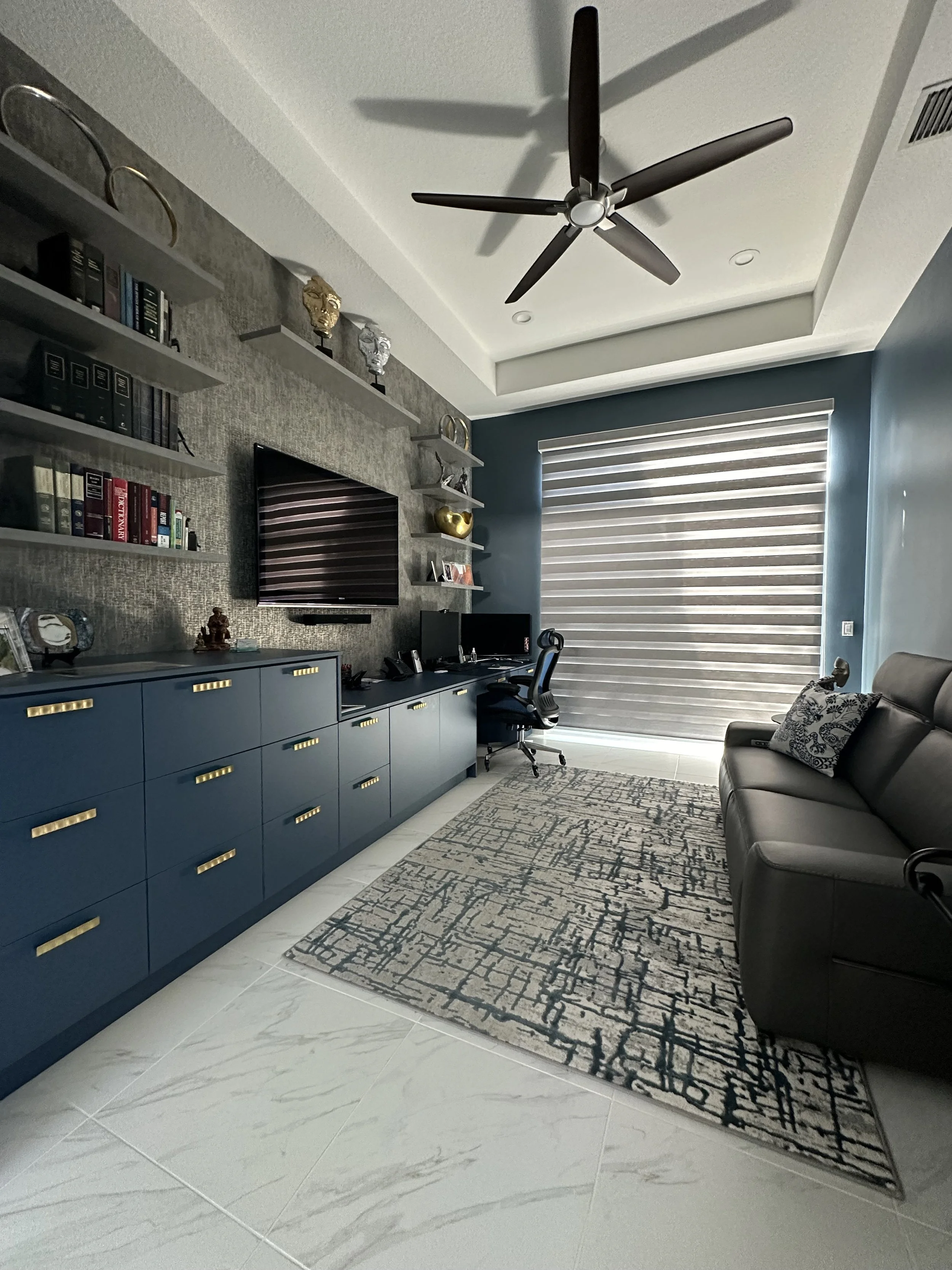 Modern home office with blue cabinetry, a black leather sofa, a patterned rug, and a computer desk near a large window with horizontal blinds. Shelves with books and decorative items on the wall, ceiling fan, and marble tile flooring.