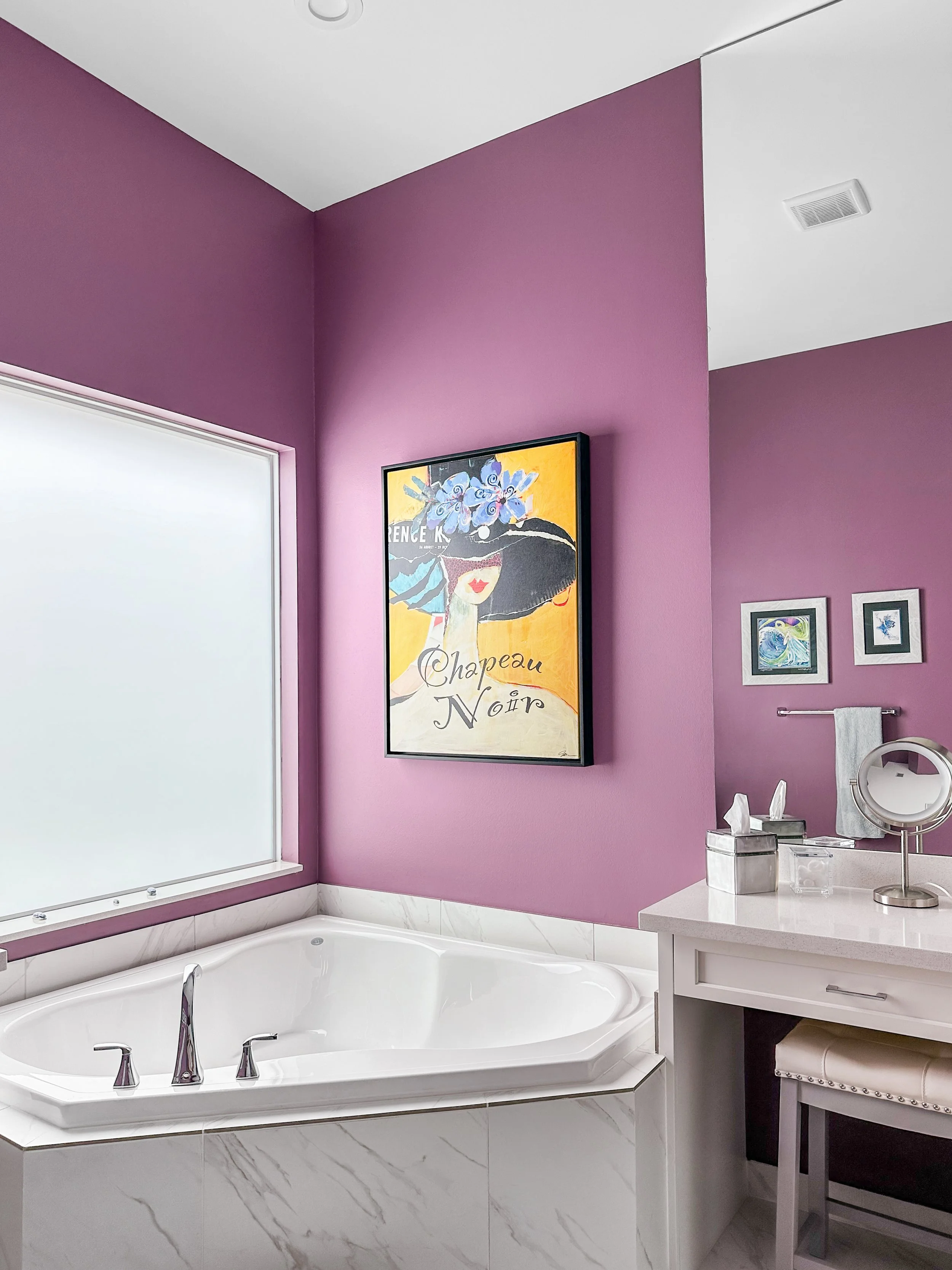 A bathroom with purple walls, a large white bathtub, a vanity with a mirror, tissue box, and framed artwork on the walls.