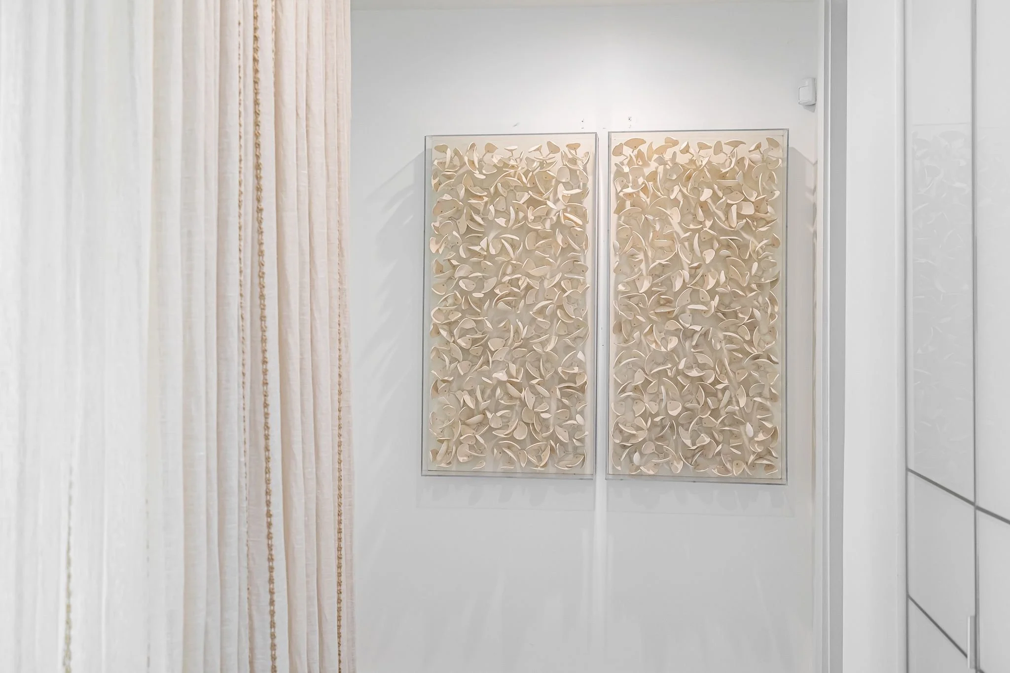 Two beige abstract textured wall art pieces hanging on a white wall, with beige drapes on the left side of the image.