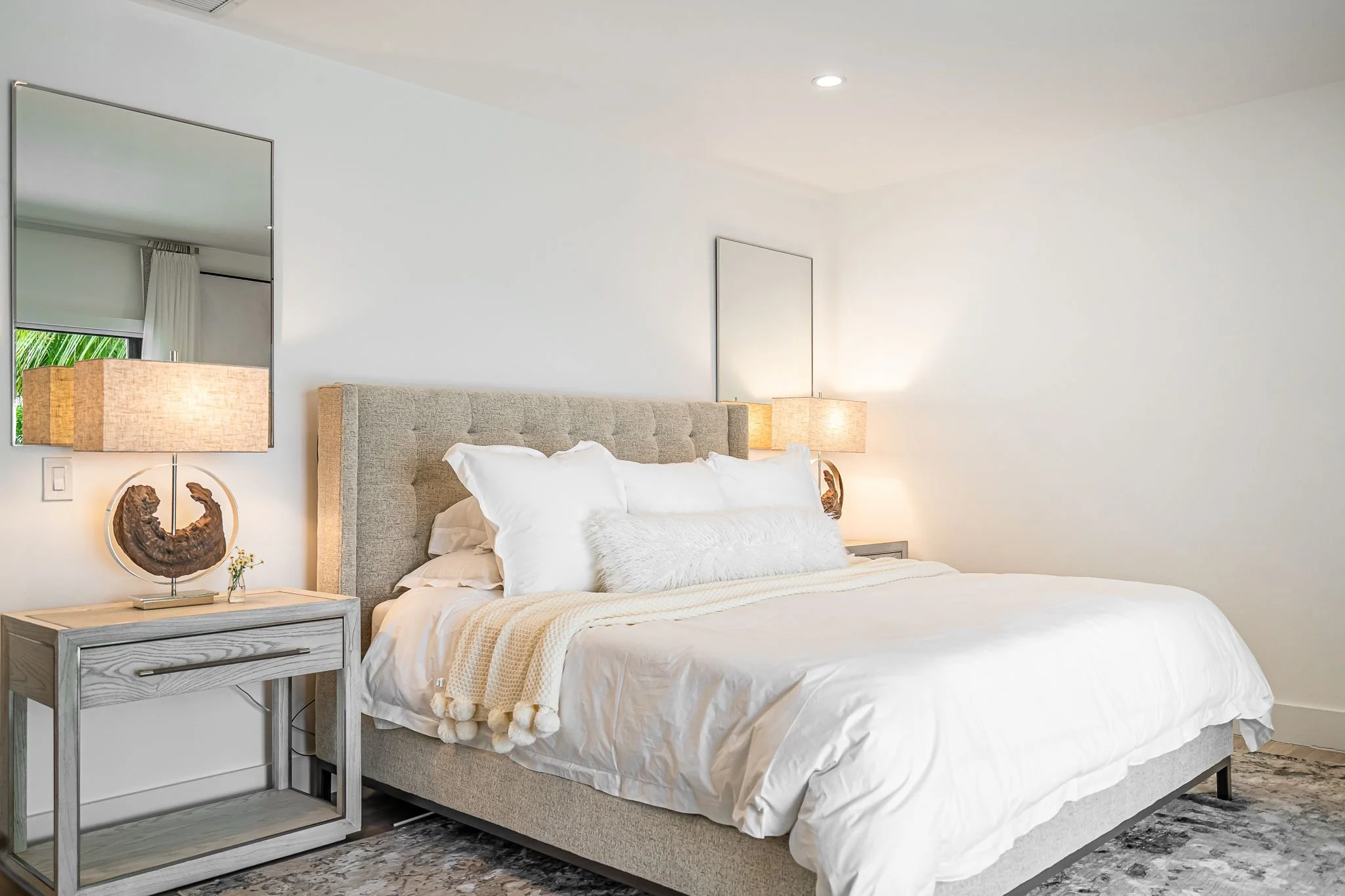 A neatly made bed with white bedding and multiple pillows, a beige upholstered headboard, two side tables with lamps, and mirrors on the wall in a modern, minimalist bedroom.