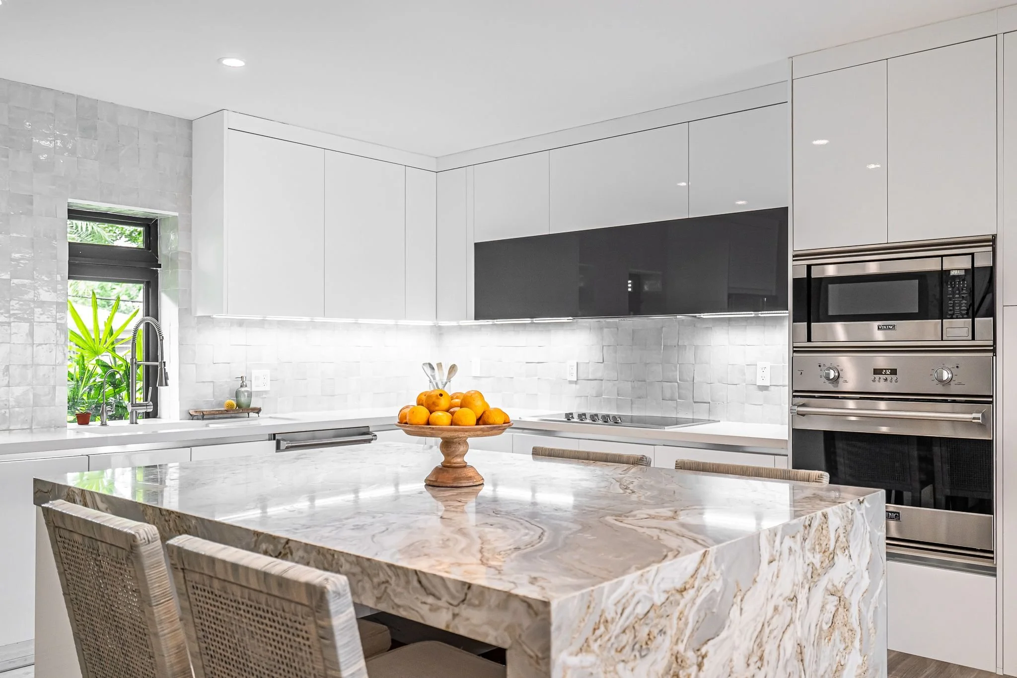 Modern kitchen with white cabinets, a marble countertop island, built-in stainless steel microwave and oven, a large window with greenery outside, and a bowl of oranges on the island.