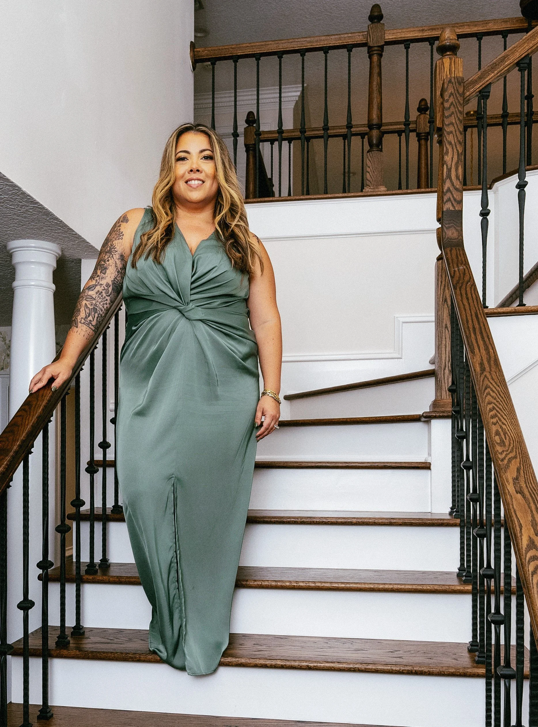 A woman in a green satin gown standing on a staircase with wooden handrails, smiling at the camera.