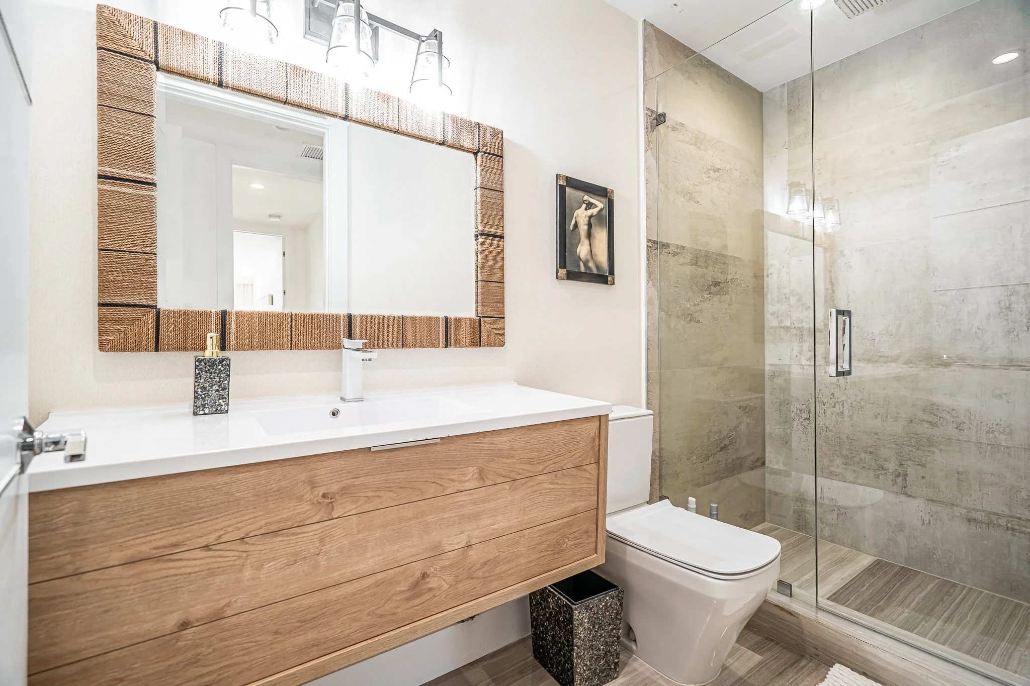 Modern bathroom with wooden vanity, large mirror, toilet, and glass shower enclosure with beige stone walls.