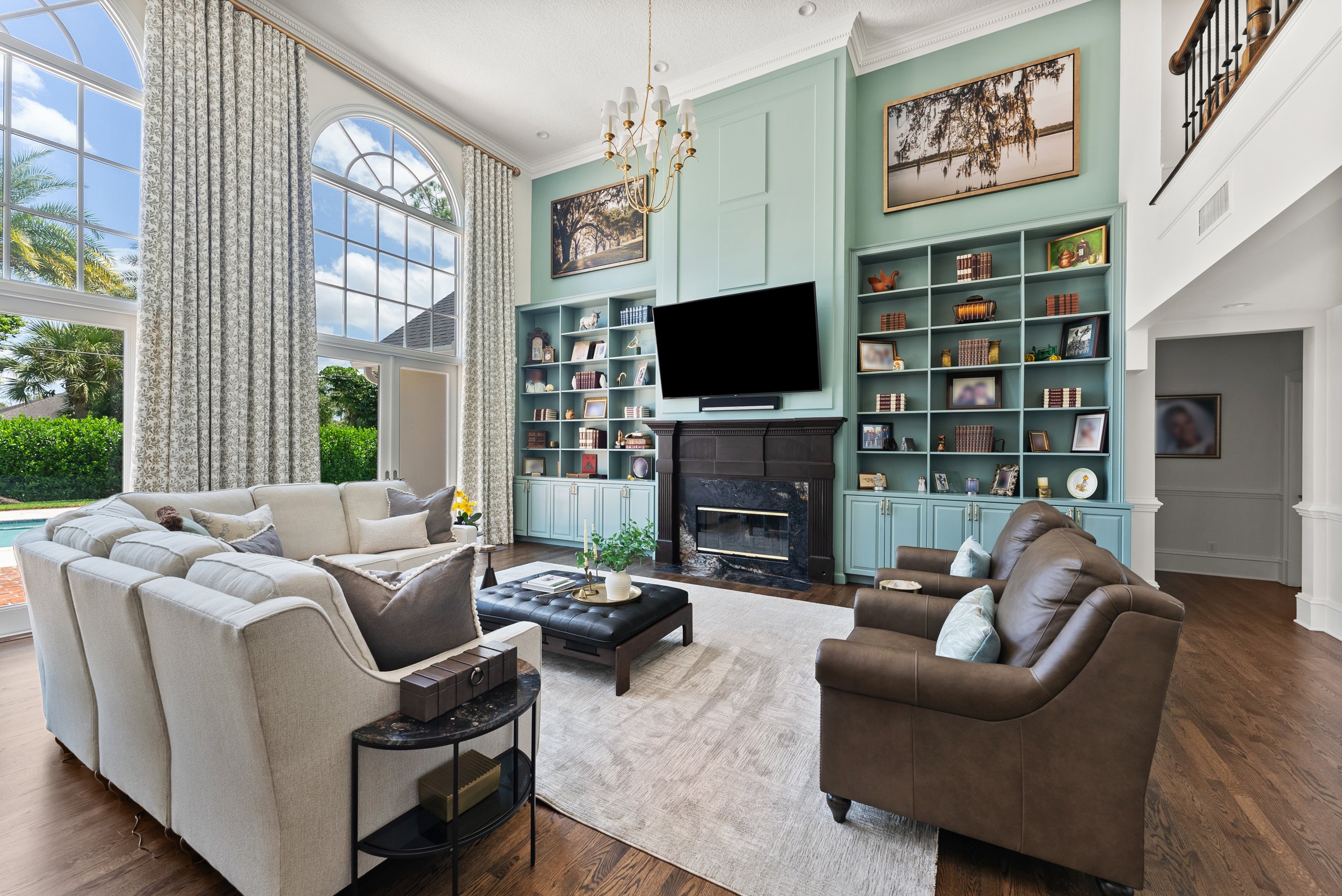 Living room with large arched windows, a cream sofa, two brown leather armchairs, a black fireplace with a TV mounted above, and built-in bookshelves filled with books and decorative items.
