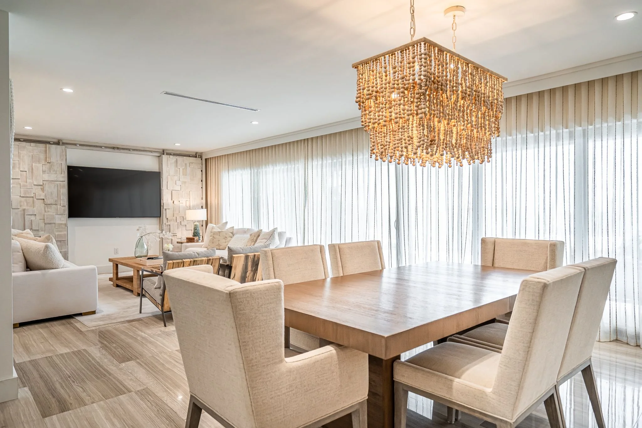 Bright living and dining room with beige chairs around a wooden table, a chandelier made of beads hanging above, beige sofas, a wall-mounted TV, and large windows with sheer curtains.