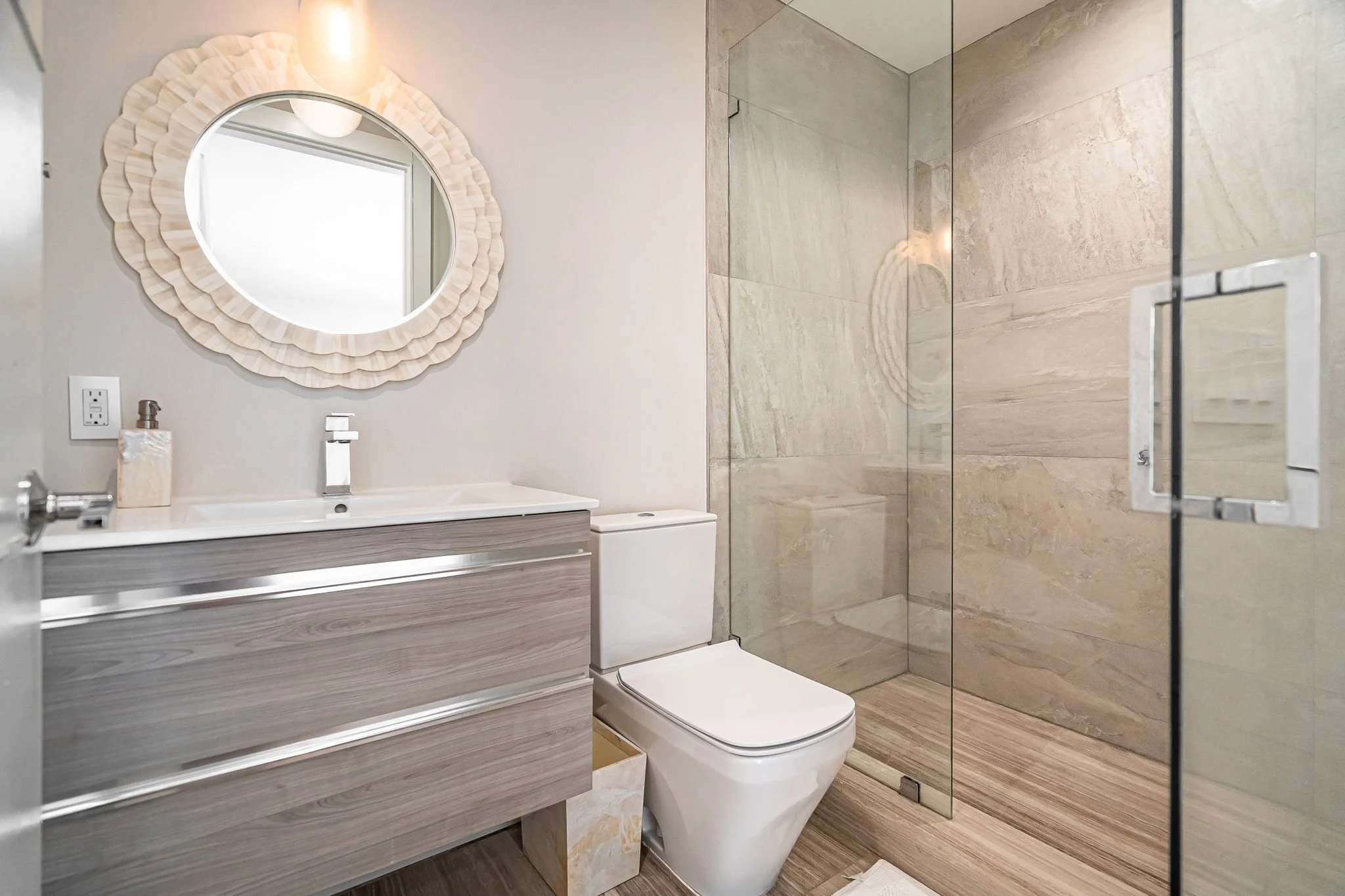 Modern bathroom with a large mirror, white sink, grey wood vanity, toilet, and a glass-enclosed walk-in shower with beige tiles.