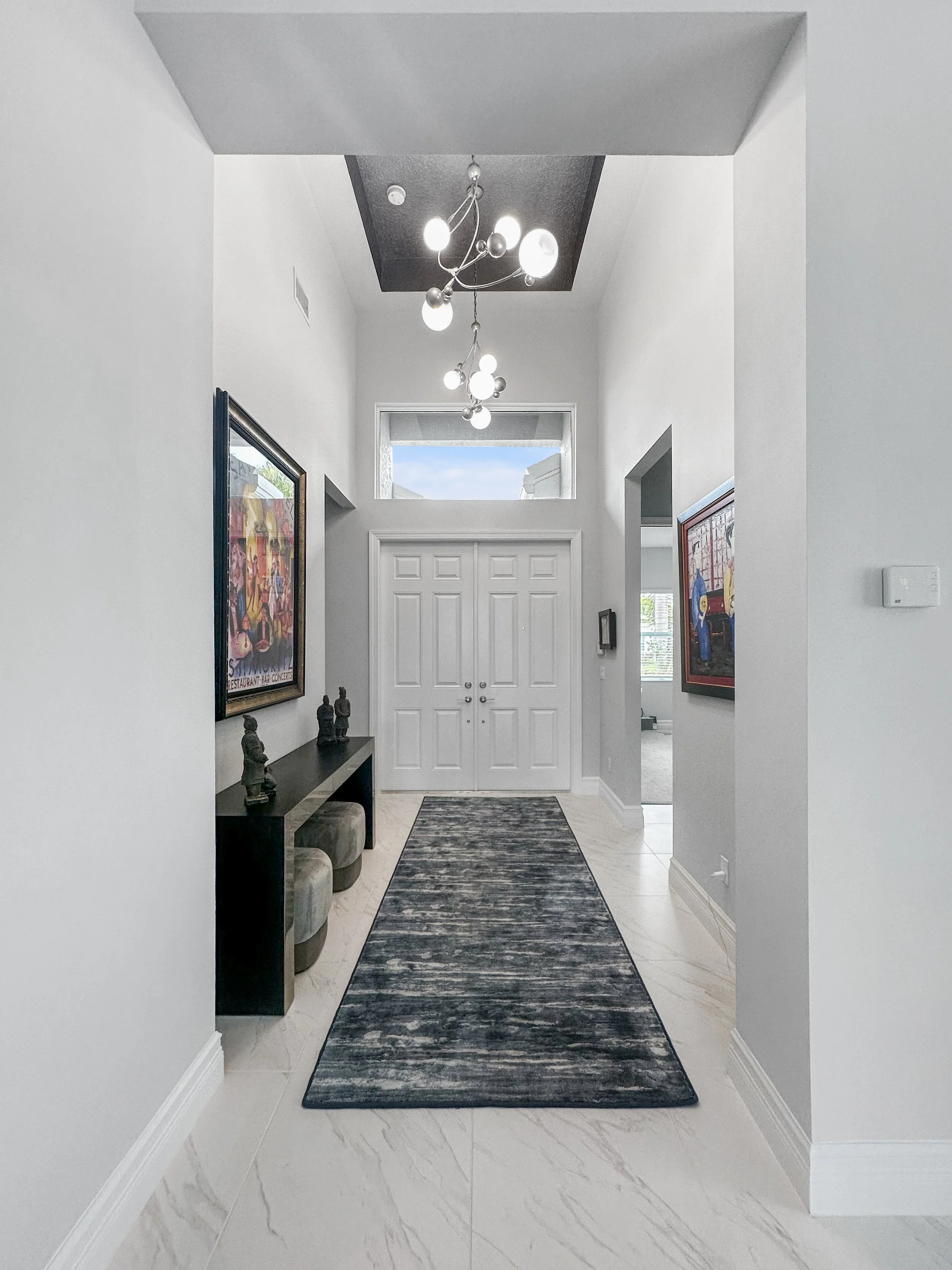 Modern entryway with a long gray rug, white walls, black console table, decorative sculptures, framed art on walls, a contemporary chandelier, double white doors, and a small window above the doors.