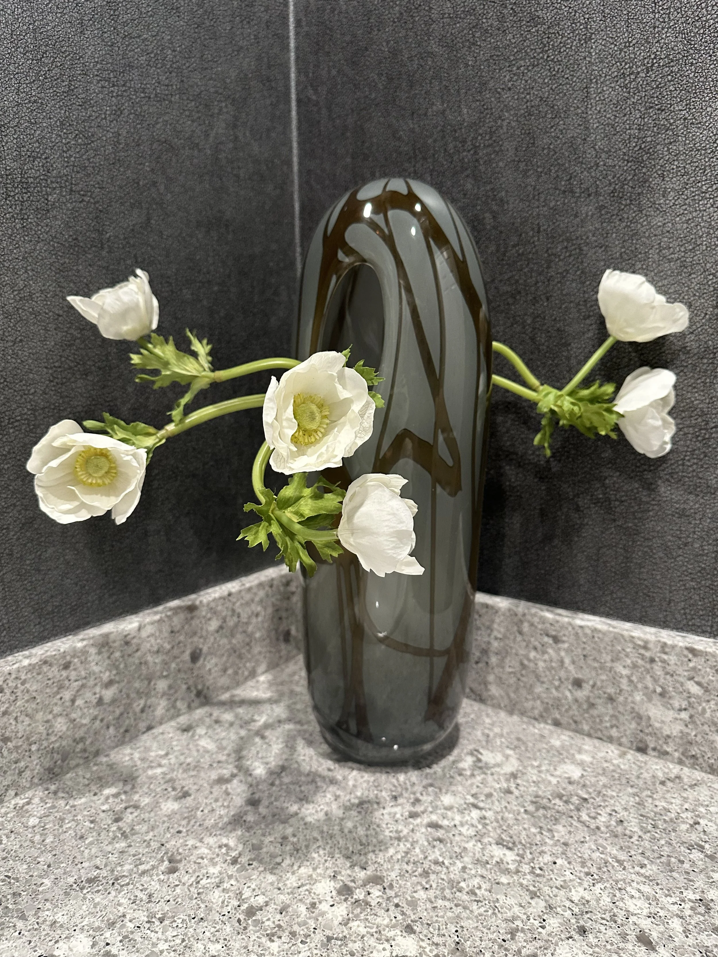 A tall, dark gray glass vase with black swirls, holding white flowers with green stems, placed on a speckled gray countertop against a dark textured wall.