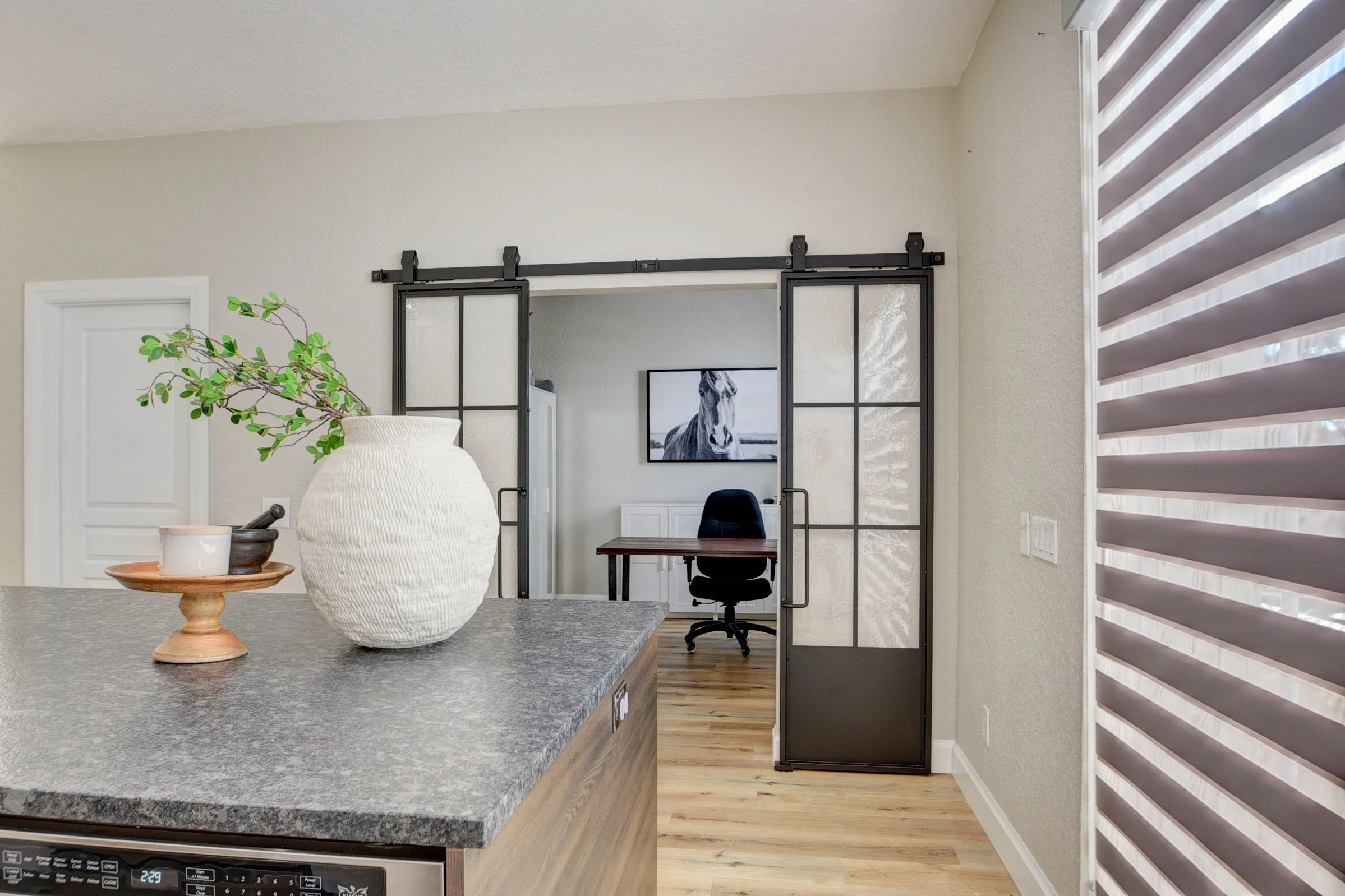 Modern kitchen with a large gray countertop, a white textured vase with green branches, and a small wooden tray with a white cup and dark mortar and pestle in the foreground. Beyond is a home office area with a black chair, a desk, a picture of a hor