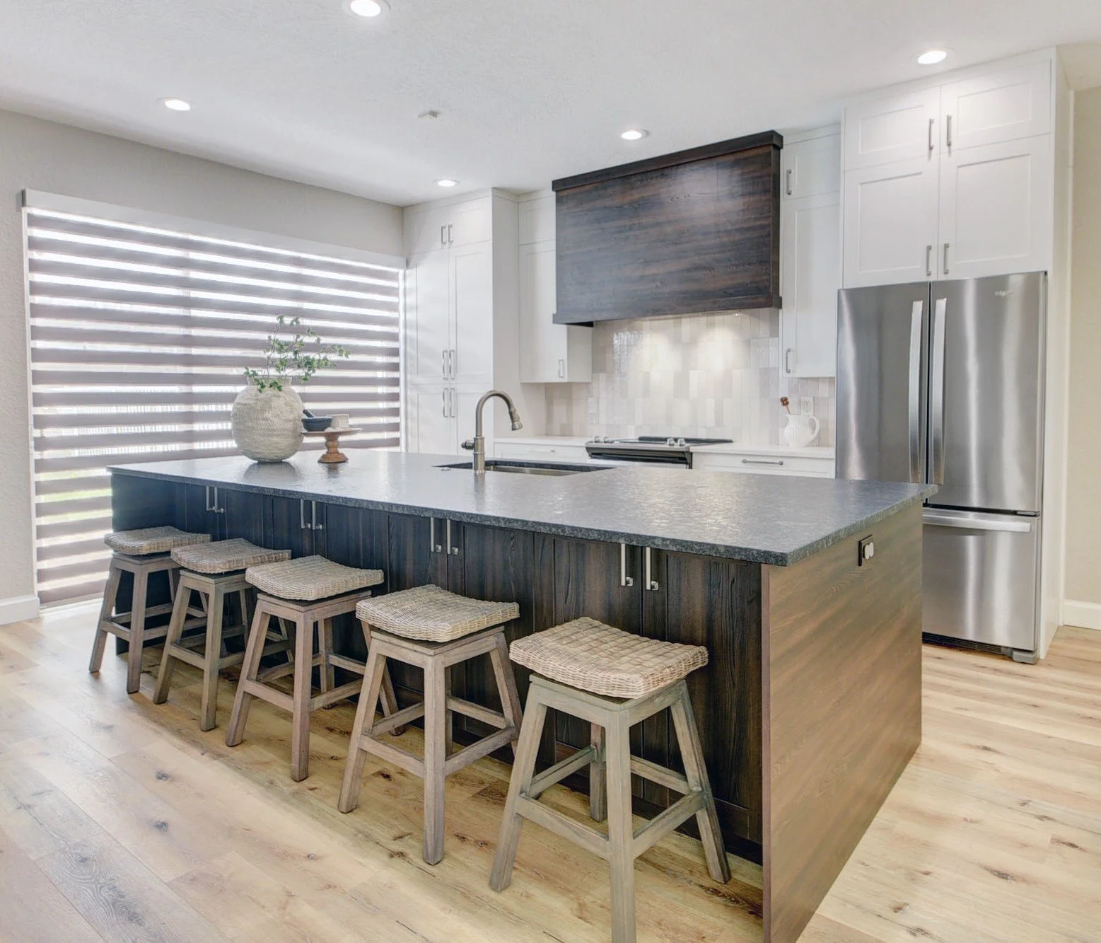 Modern kitchen with dark wood island, light hardwood flooring, white cabinets, and stainless steel refrigerator, with large window with horizontal blinds.