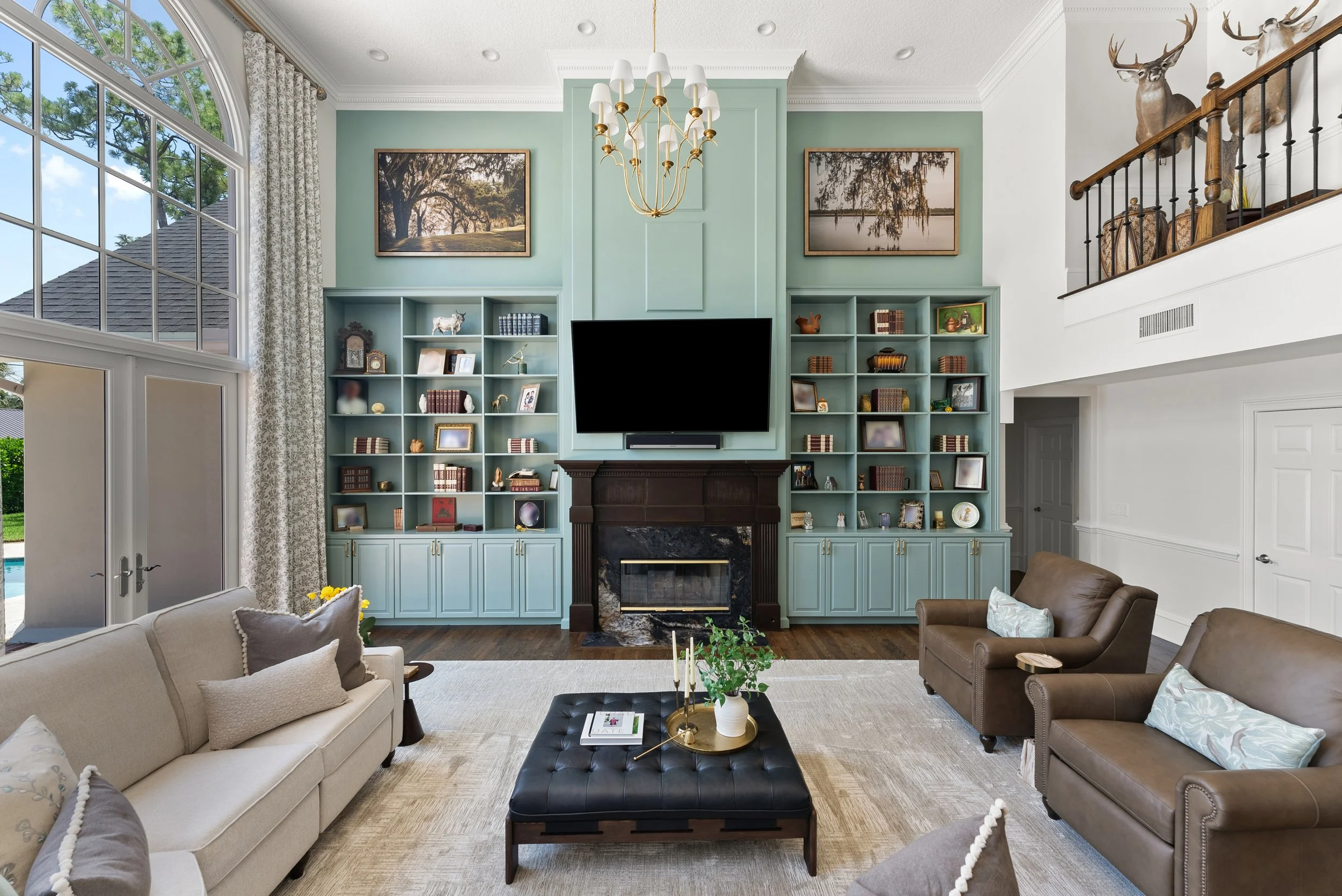 Living room with a large window, built-in shelves with books and decor, a black fireplace, a mounted TV, two velvet armchairs, a beige sofa, a black ottoman with a tray and plant, and mounted animal heads on the upper railing.