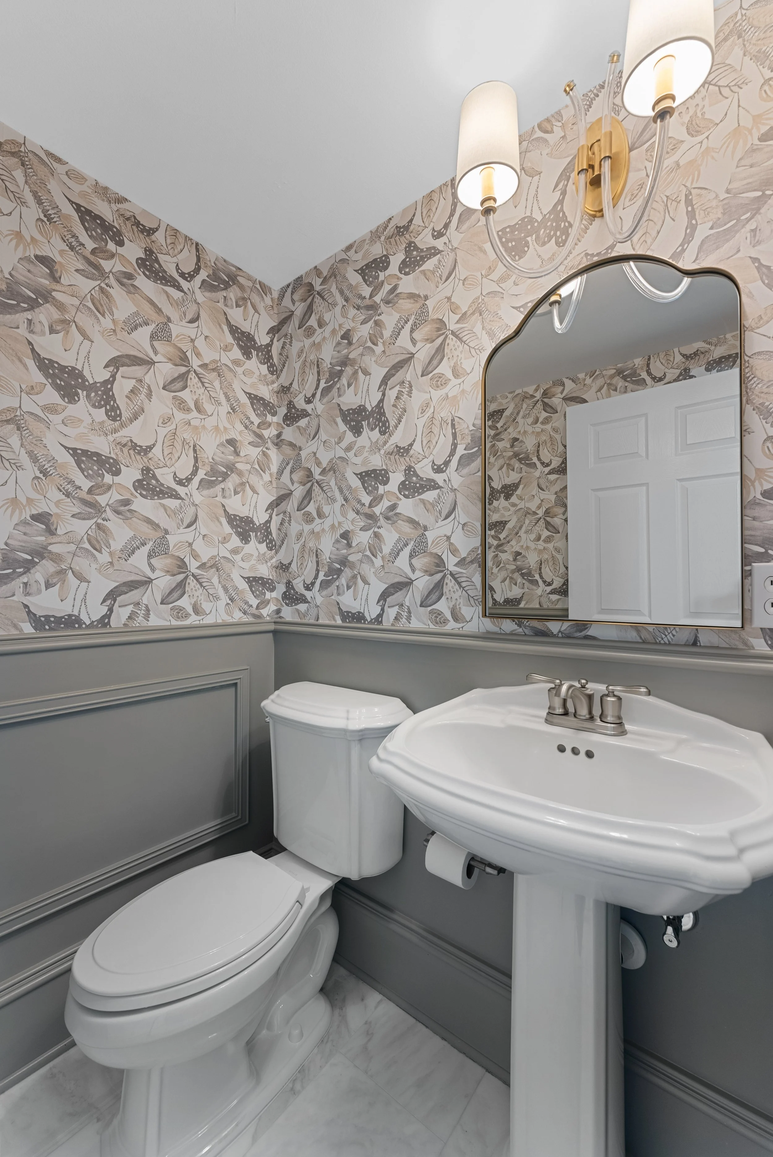 Bathroom with wallpaper featuring butterfly and leaf patterns, white toilet, white pedestal sink, oval mirror with gold trim, and wall-mounted light fixture with two lights.