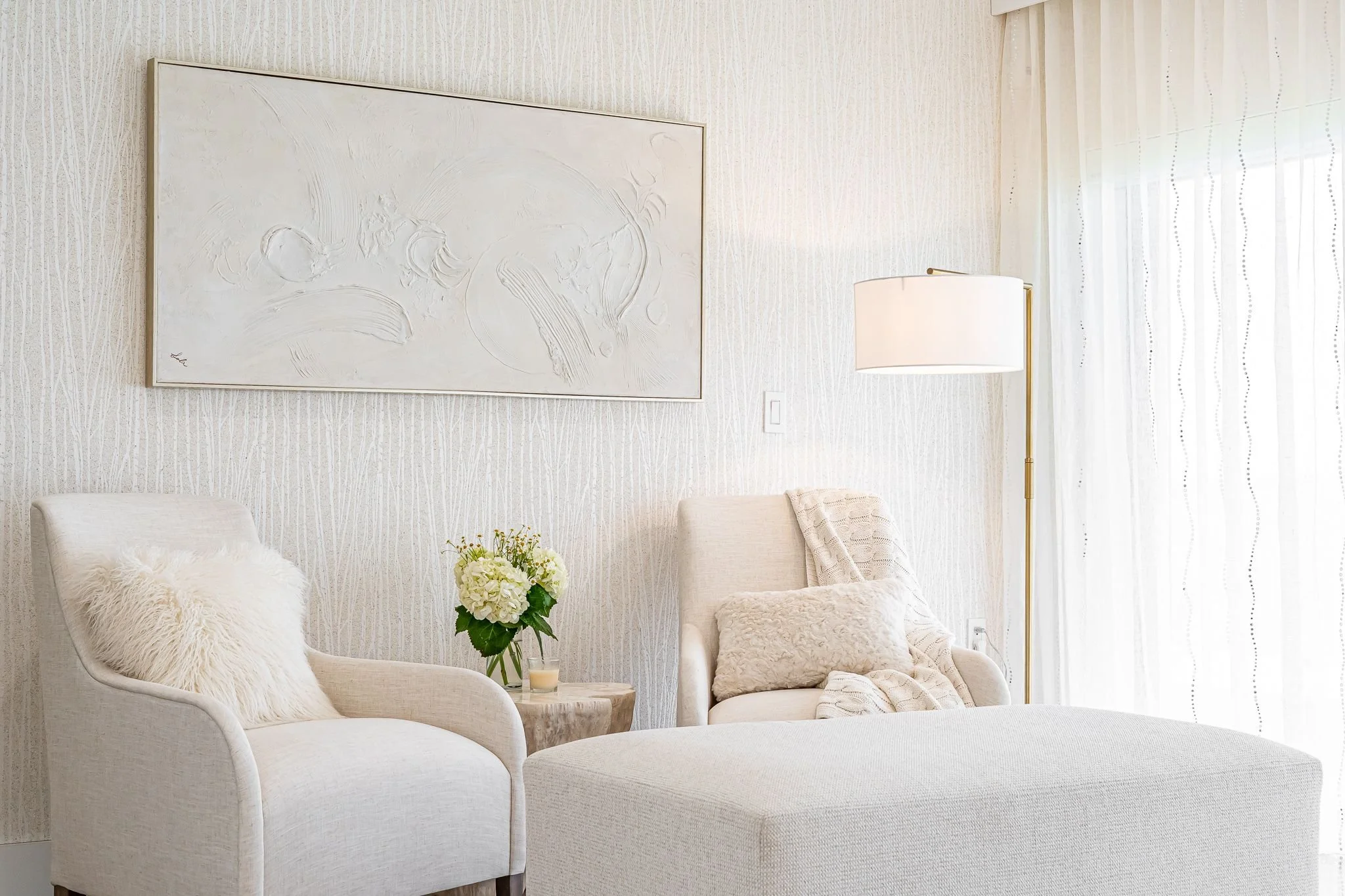 A living room with white armchairs, a floral arrangement, a large abstract painting, a floor lamp, sheer curtains, and textured walls.