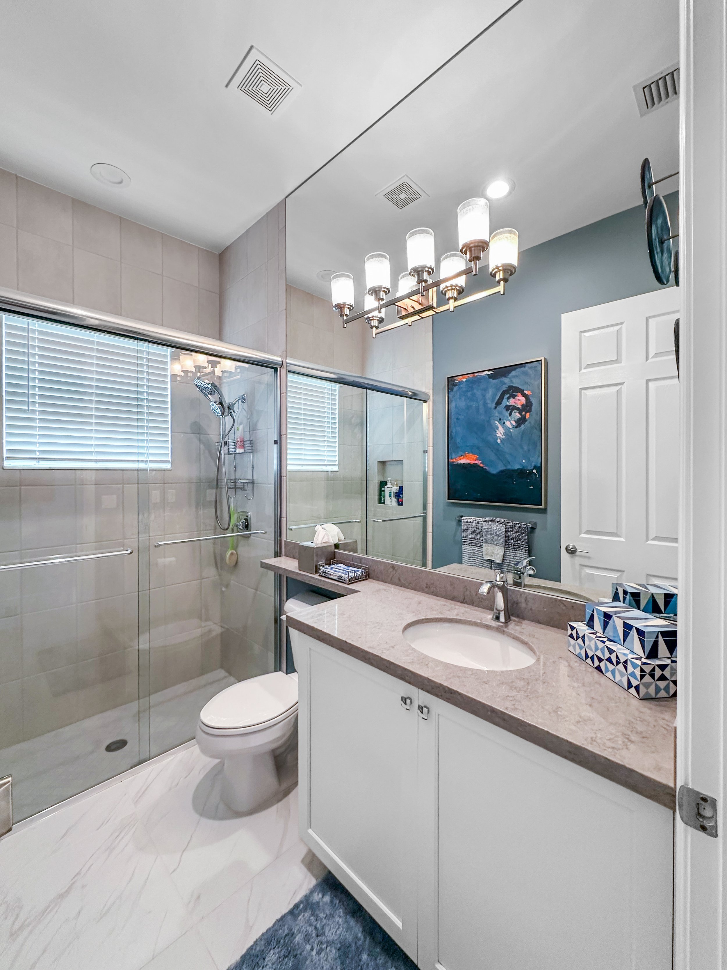 A modern bathroom with a white vanity with a beige countertop, a sink with a silver faucet, and a large mirror. There is a colorful abstract painting on the blue wall, a towel hanging on a rack, and a framed set of tissue boxes on the countertop. A g