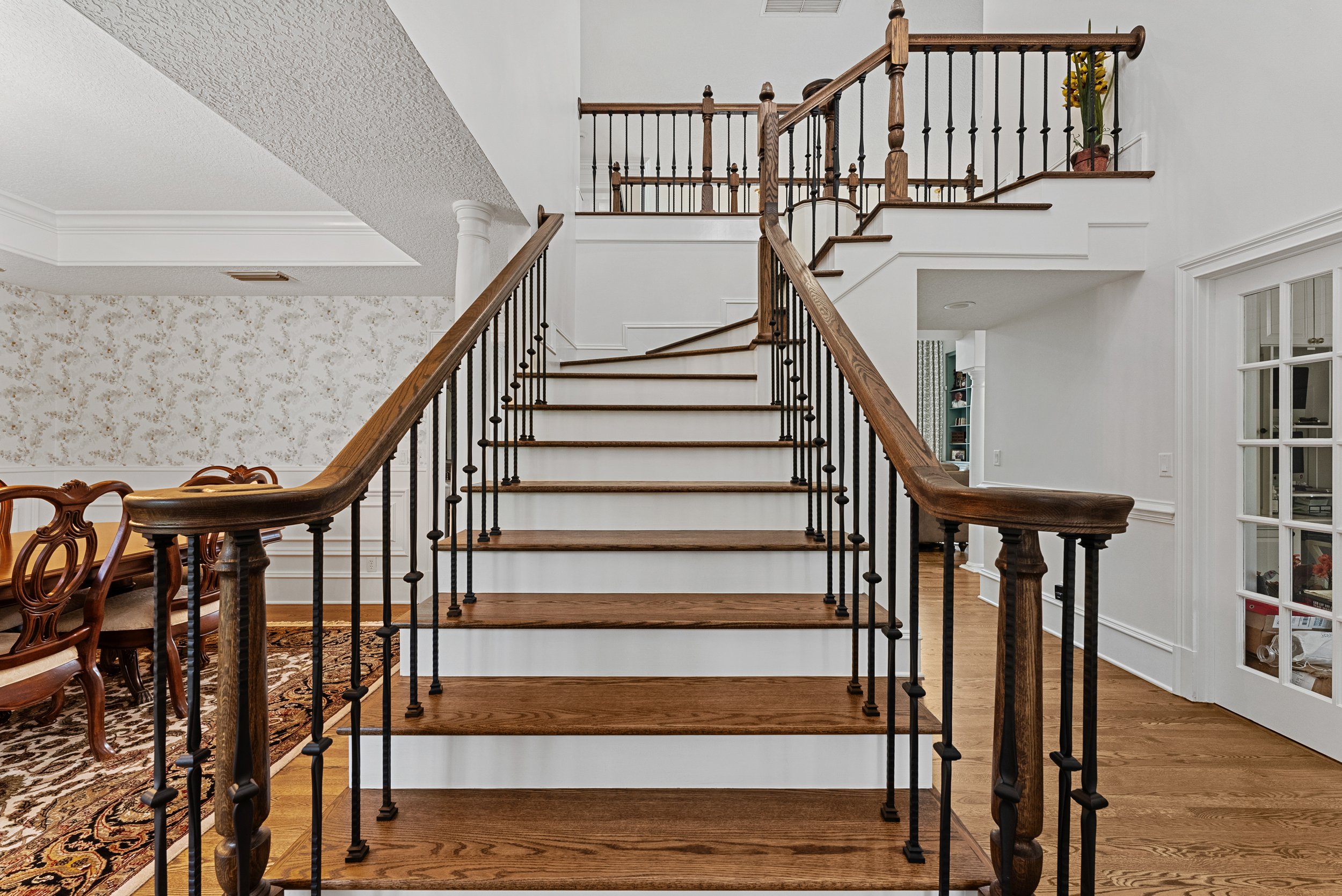 Wood staircase with black metal balusters in a house interior, leading to a second floor with a wooden handrail, surrounded by white walls and a partial view of a dining area with chairs and a rug.