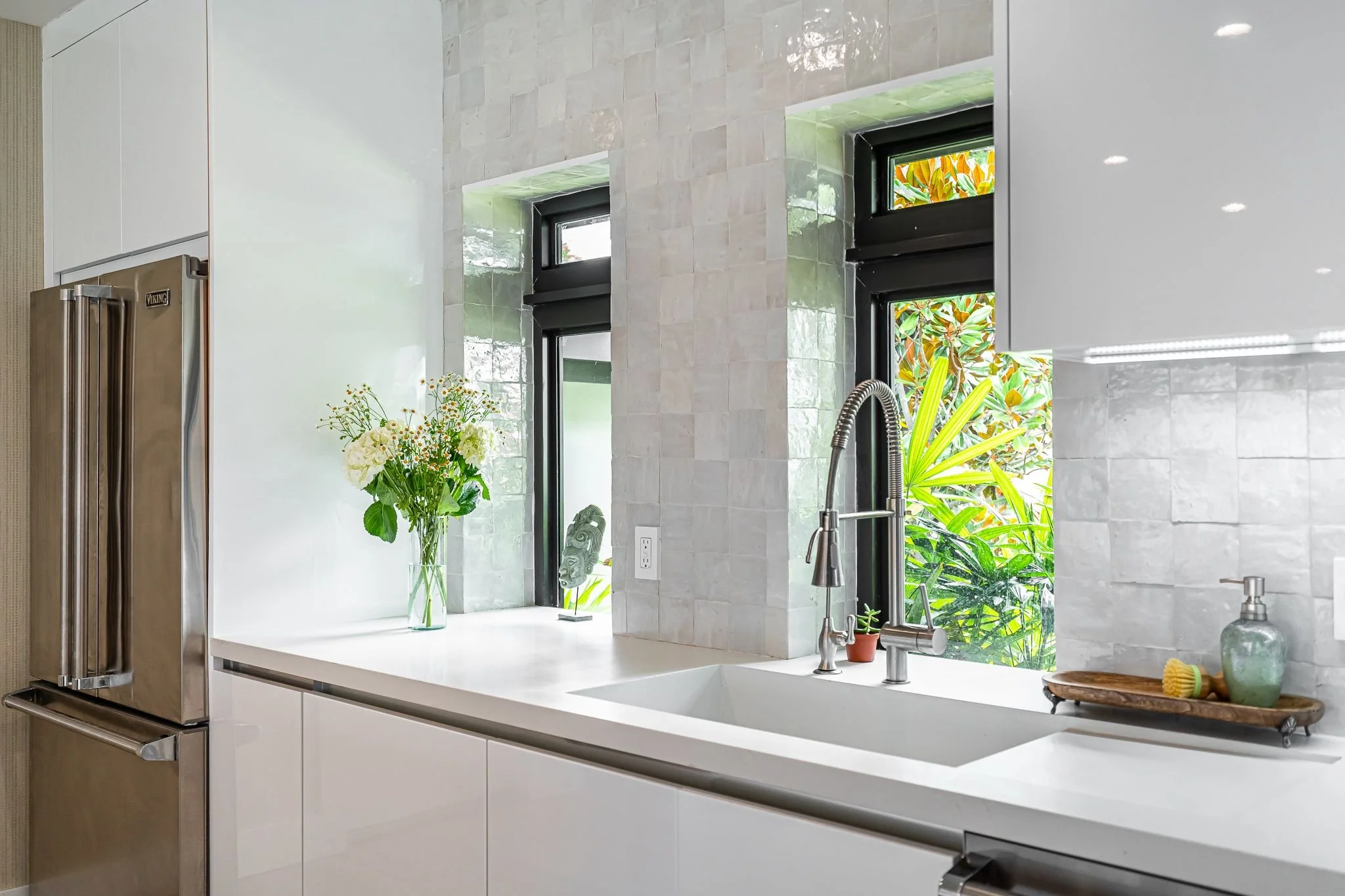 Modern kitchen with white counters, a stainless steel refrigerator, a black framed window showing green foliage outside, a vase with white flowers on the counter, and a soap dispenser on a wooden tray.