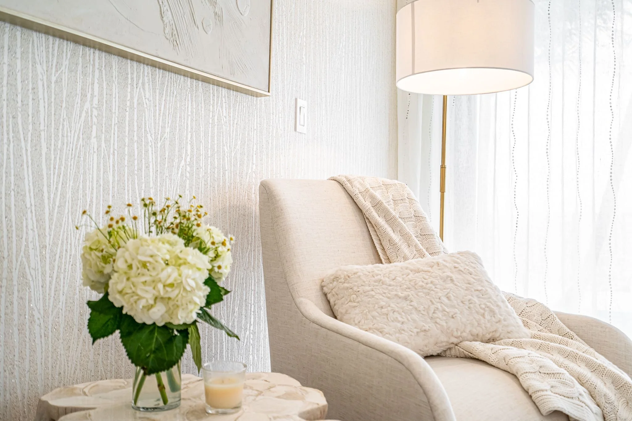A cozy, light-colored living room with a beige armchair, a knitted blanket, and a fluffy pillow. A side table with a vase of white flowers and a small candle. A tall floor lamp and sheer curtains in the background.