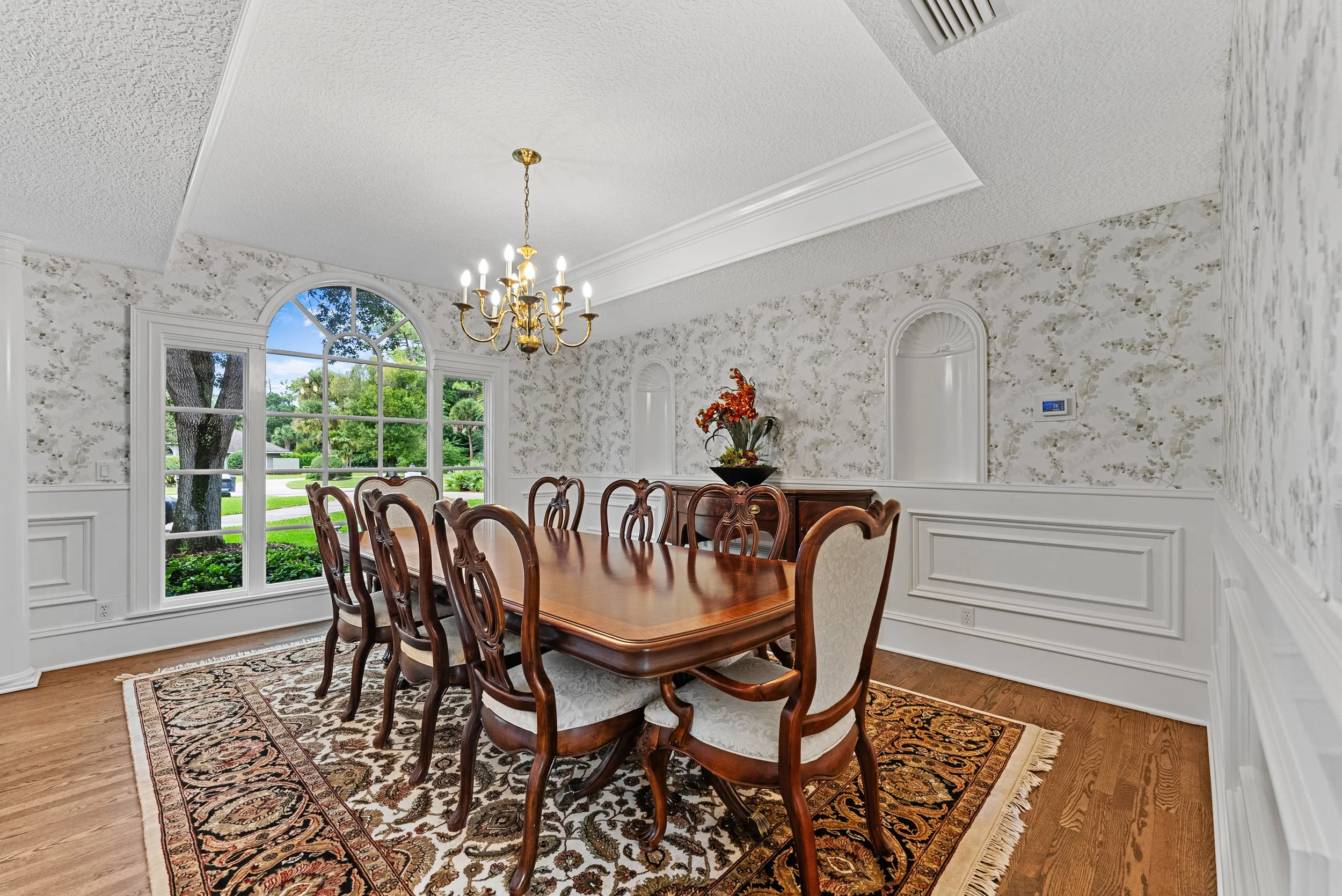 A dining room with a large wooden table, eight chairs, a floral rug, a chandelier, and a window showing a view of a lawn and trees outside.
