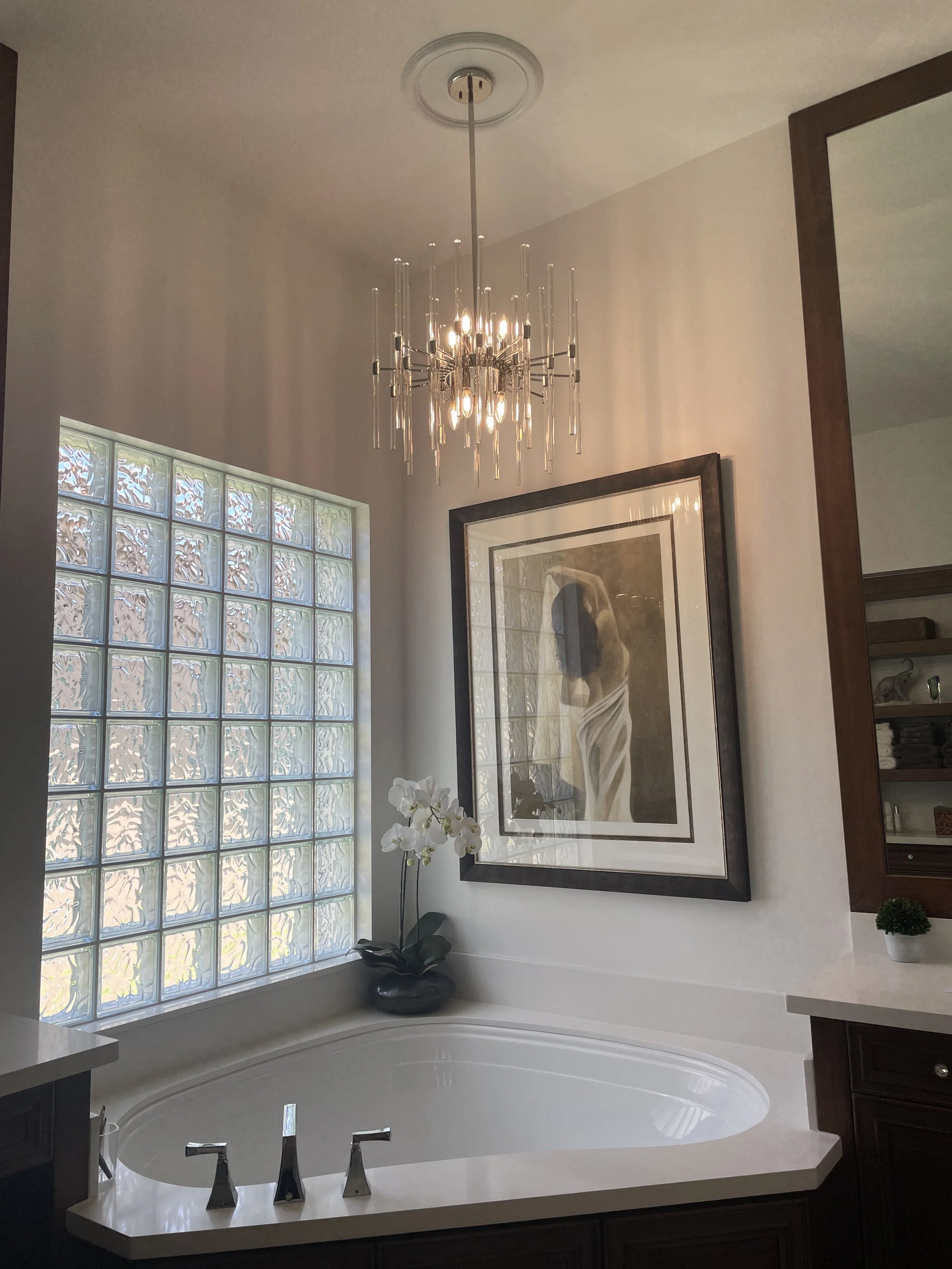 A bathroom with a corner bathtub, a large framed artwork of a woman, a glass block window, a white orchid plant in a pot, and a modern chandelier hanging from the ceiling.