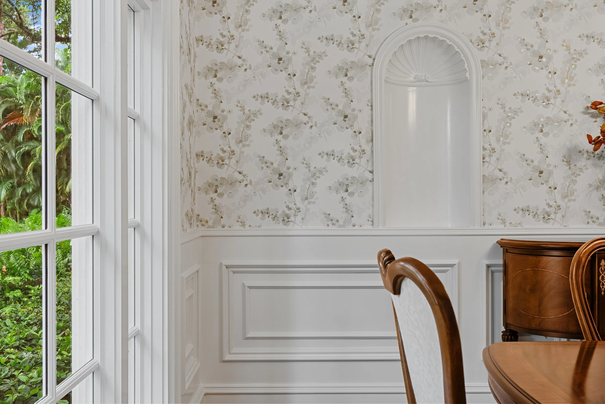 A corner of a dining room with white floral wallpaper, white wood paneling, a window with greenery outside, and part of a wooden dining table and chair.