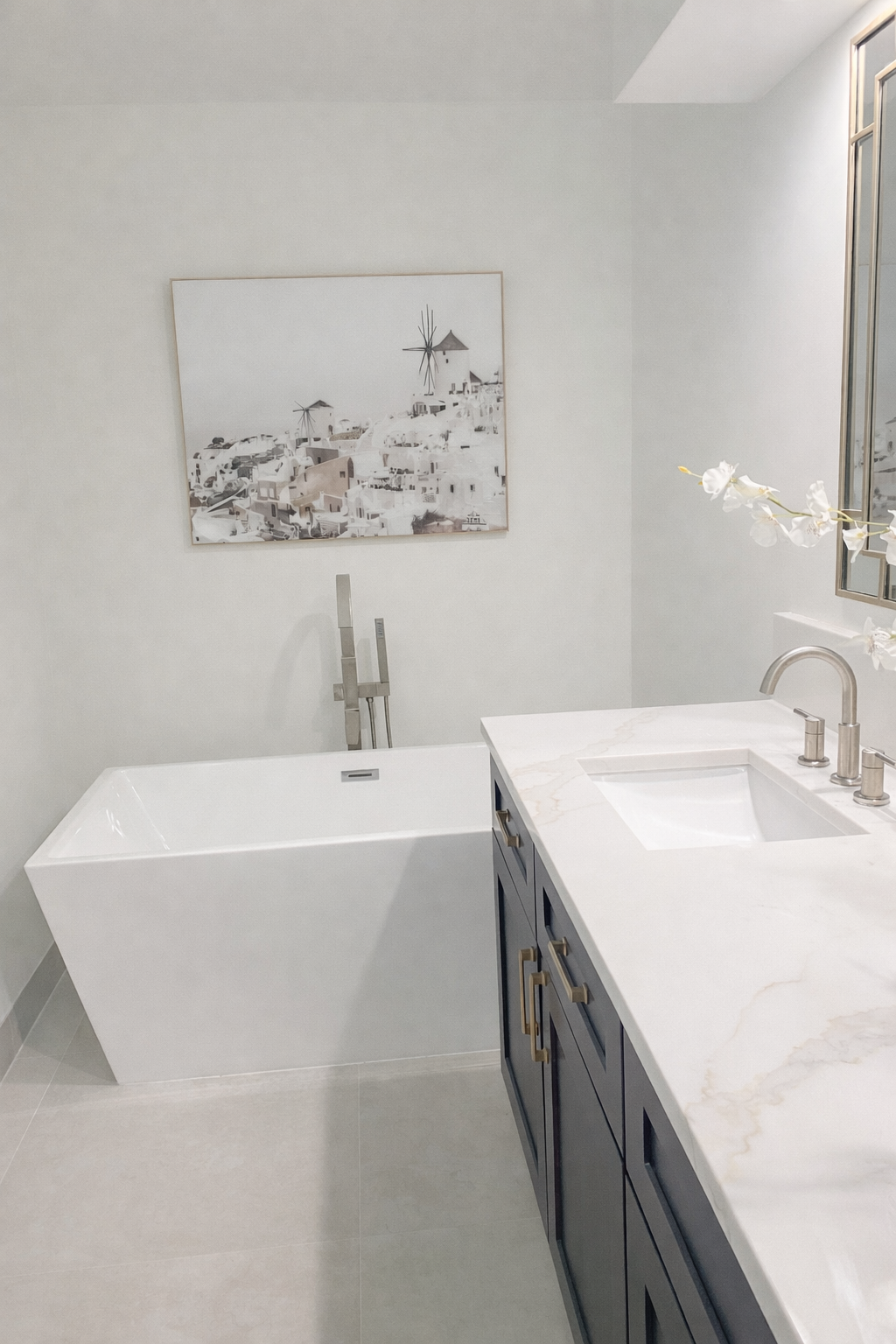 Modern bathroom with white bathtub, marble countertop with a sink, and a framed artwork of white buildings with windmills, all in neutral tones.