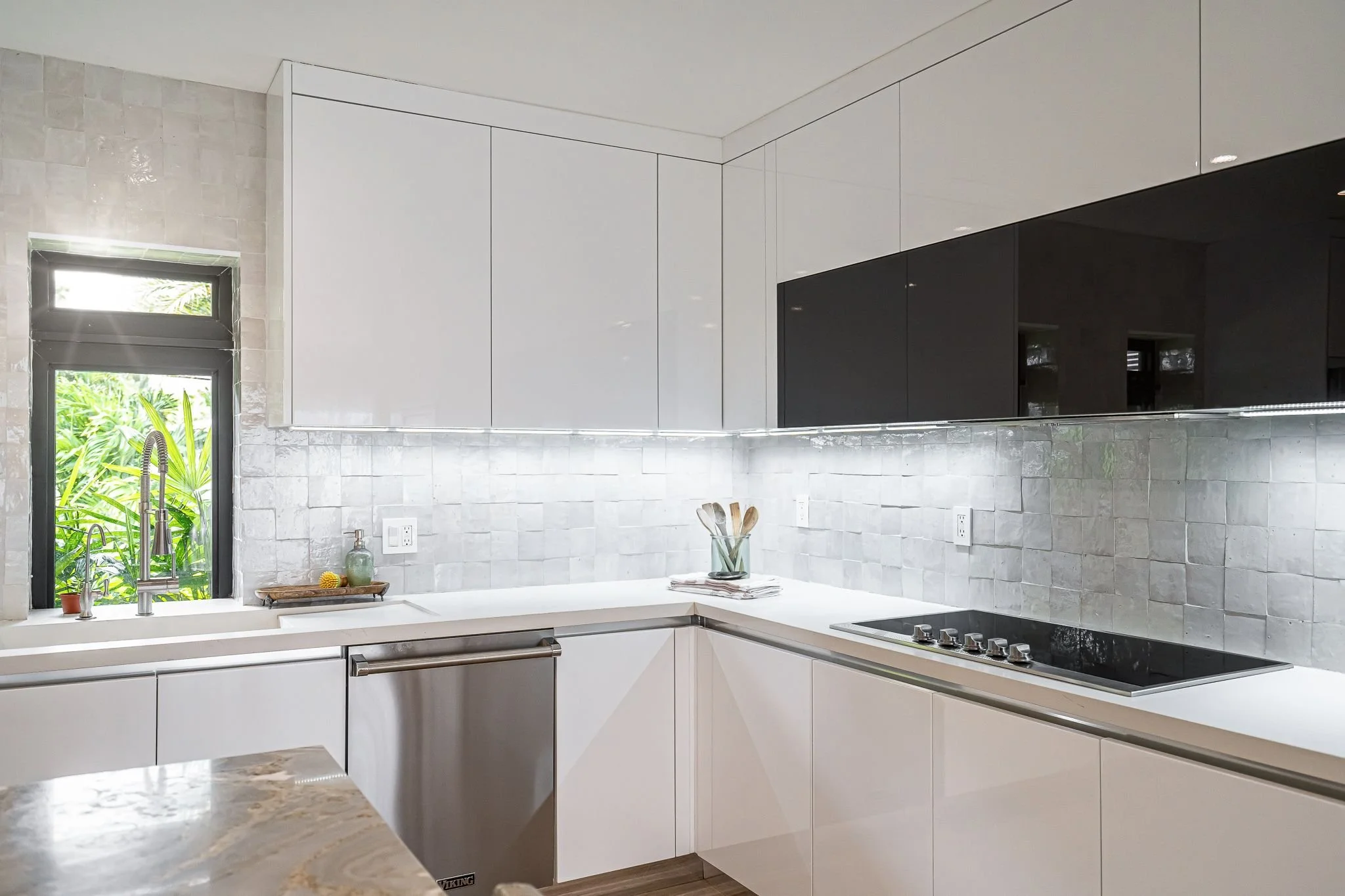 Modern kitchen with white lower cabinets, black upper cabinets, a grey tiled backsplash, a small window above the sink with greenery outside, a stove with knobs, a few utensils in a glass jar, and a window with sunlight coming through.