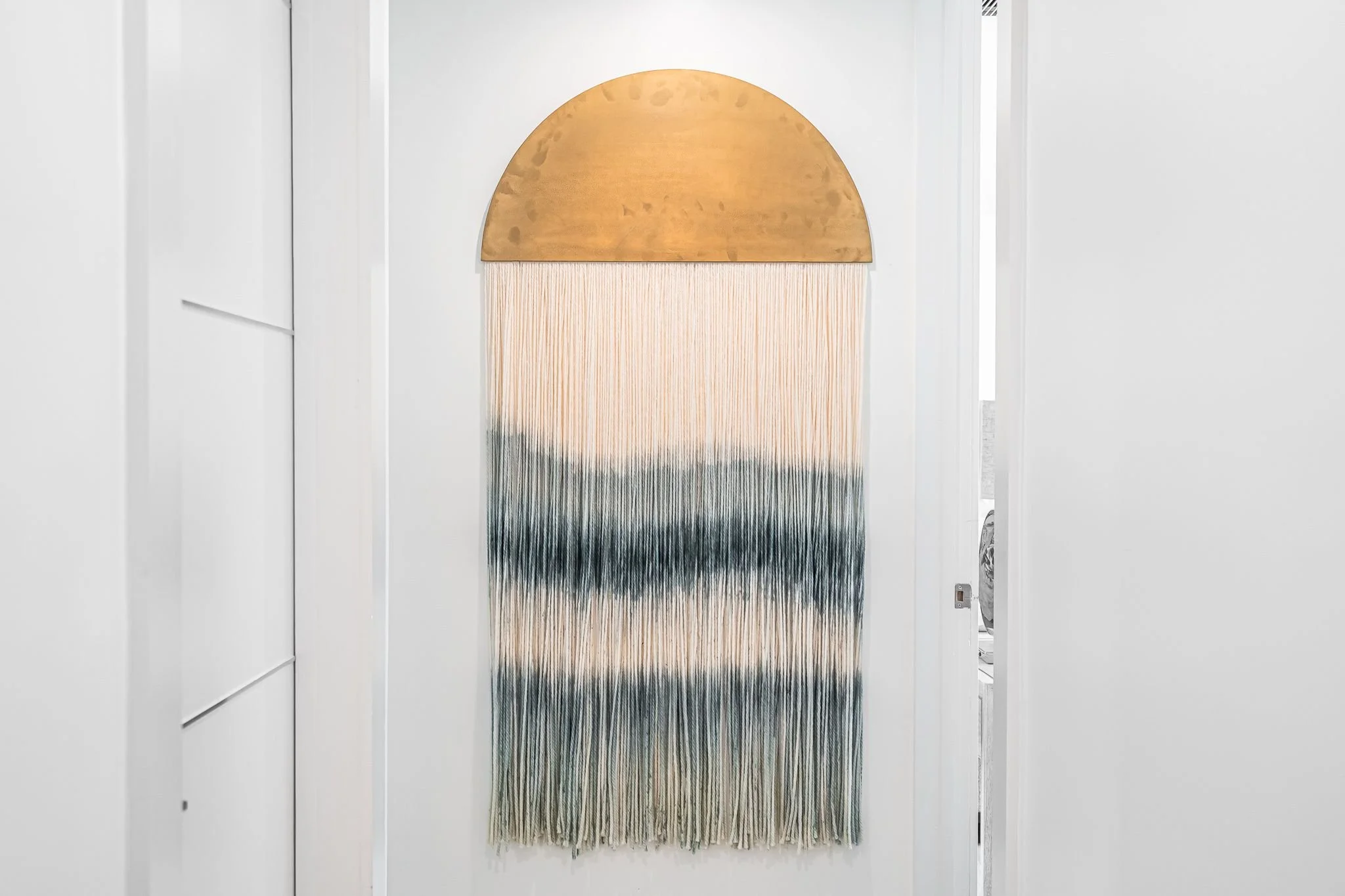 Decorative wall hanging with an arched top, wooden semi-circular shape, and woven strands in beige, white, black, and green colors, hanging on a white wall.