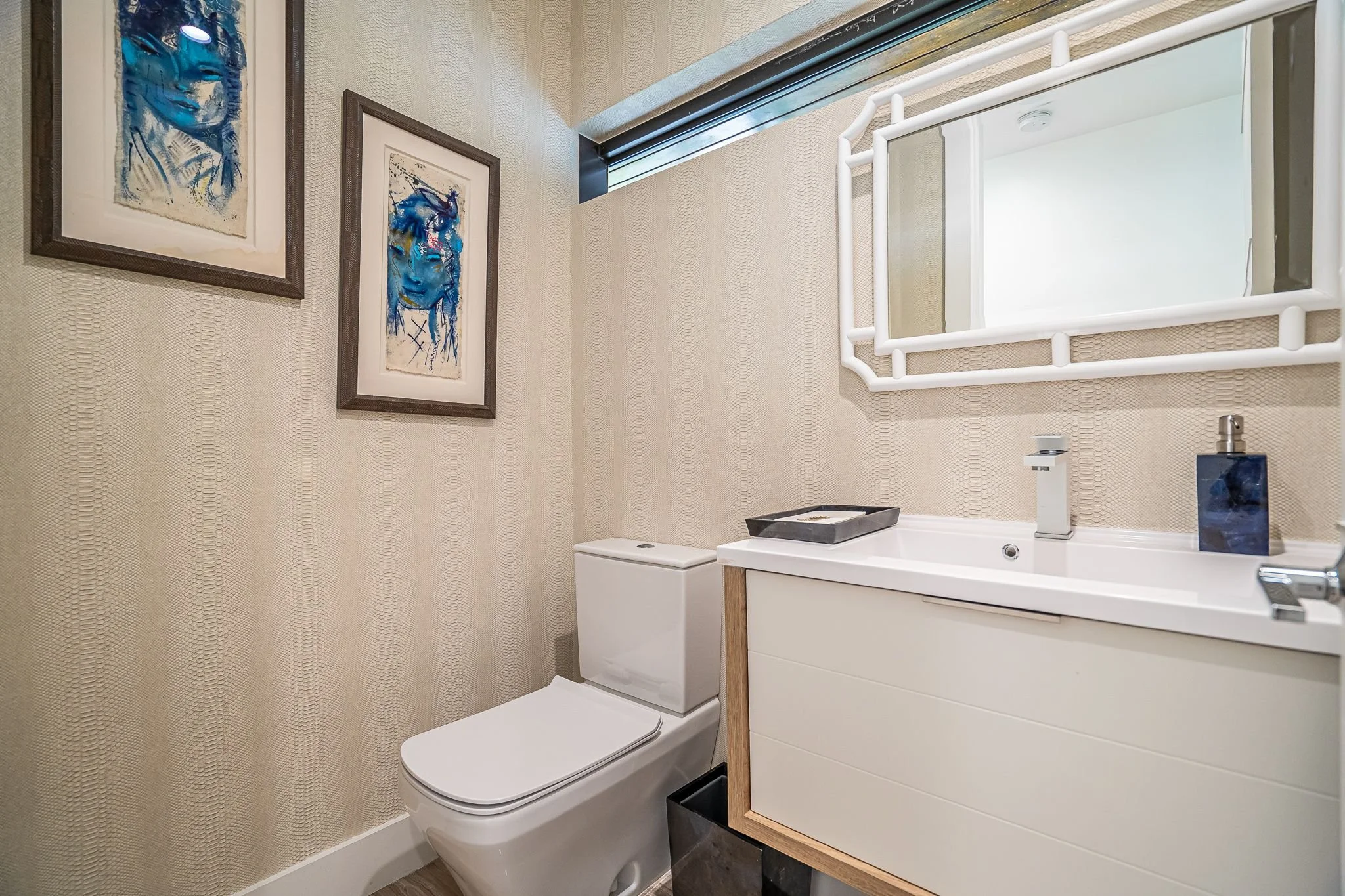 Contemporary bathroom with white toilet, white vanity with black soap dispenser, and wall art featuring blue portraits, beige textured wallpaper, and a white mirror above the sink.