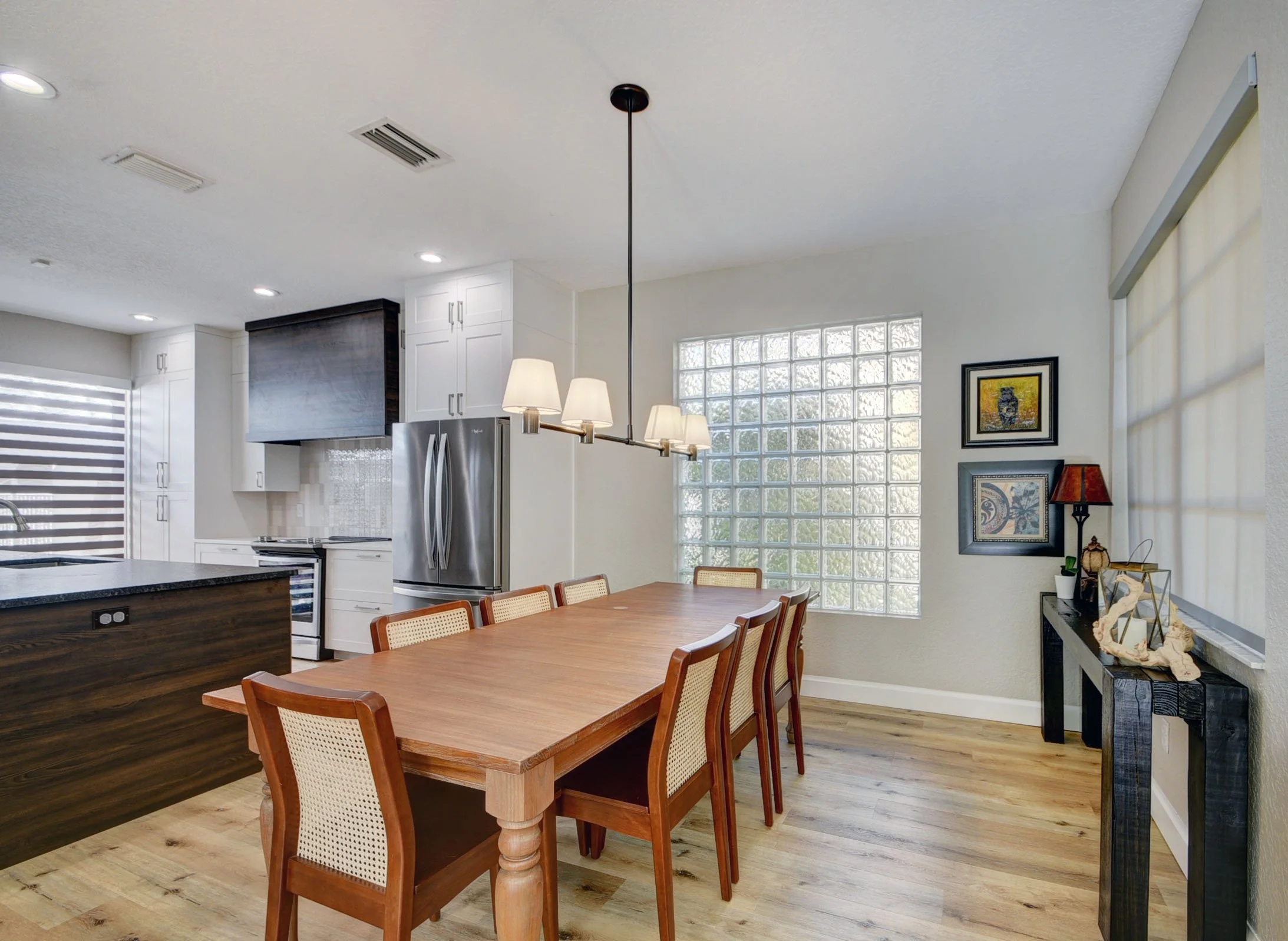 Dining area with a wooden table and six chairs, a black side table with decorations, wall art, a window with textured glass, and a kitchen in the background with white cabinets and stainless steel appliances.