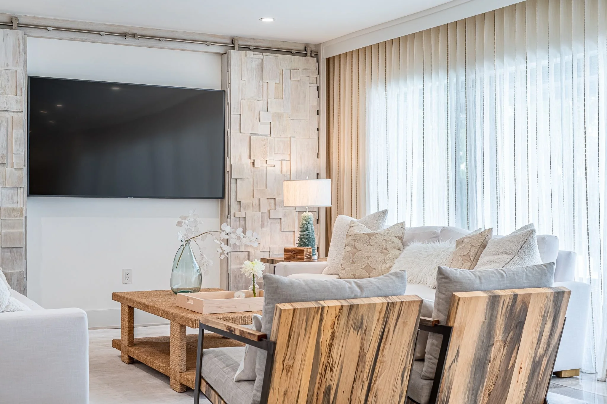Living room with white sofa, wooden armchairs, a wooden coffee table with a glass vase and flowers, a side table with a lamp and decorative tree, large windows with sheer curtains, and a mounted flat-screen TV on the wall.