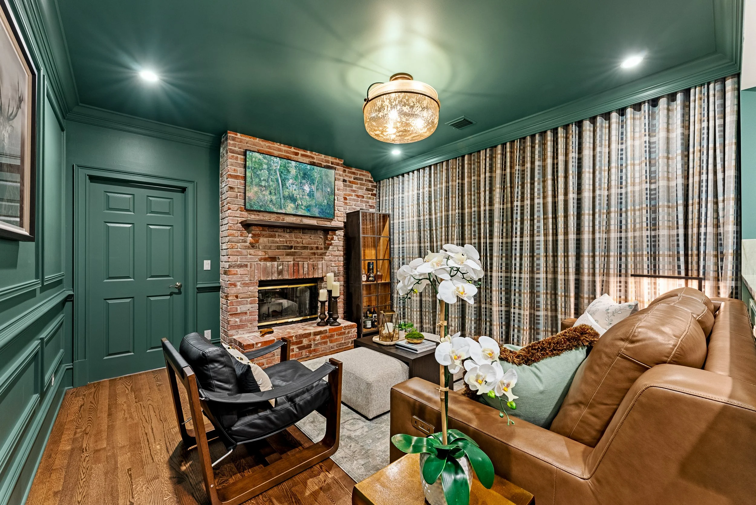 Living room with green paneled walls, brick fireplace, flat-screen TV, plaid curtains, leather sofa, black armchair, wooden floors, flower arrangement, and decorative candles.