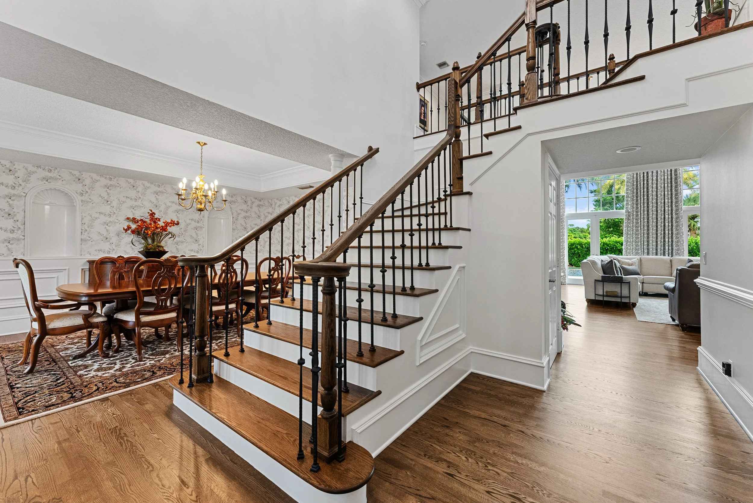 Interior view of a home showing a staircase with wooden steps and black metal balusters, leading upstairs. To the left, a dining area with a wooden table, chairs, and a chandelier, decorated with a vase of orange flowers. To the right, a living room 