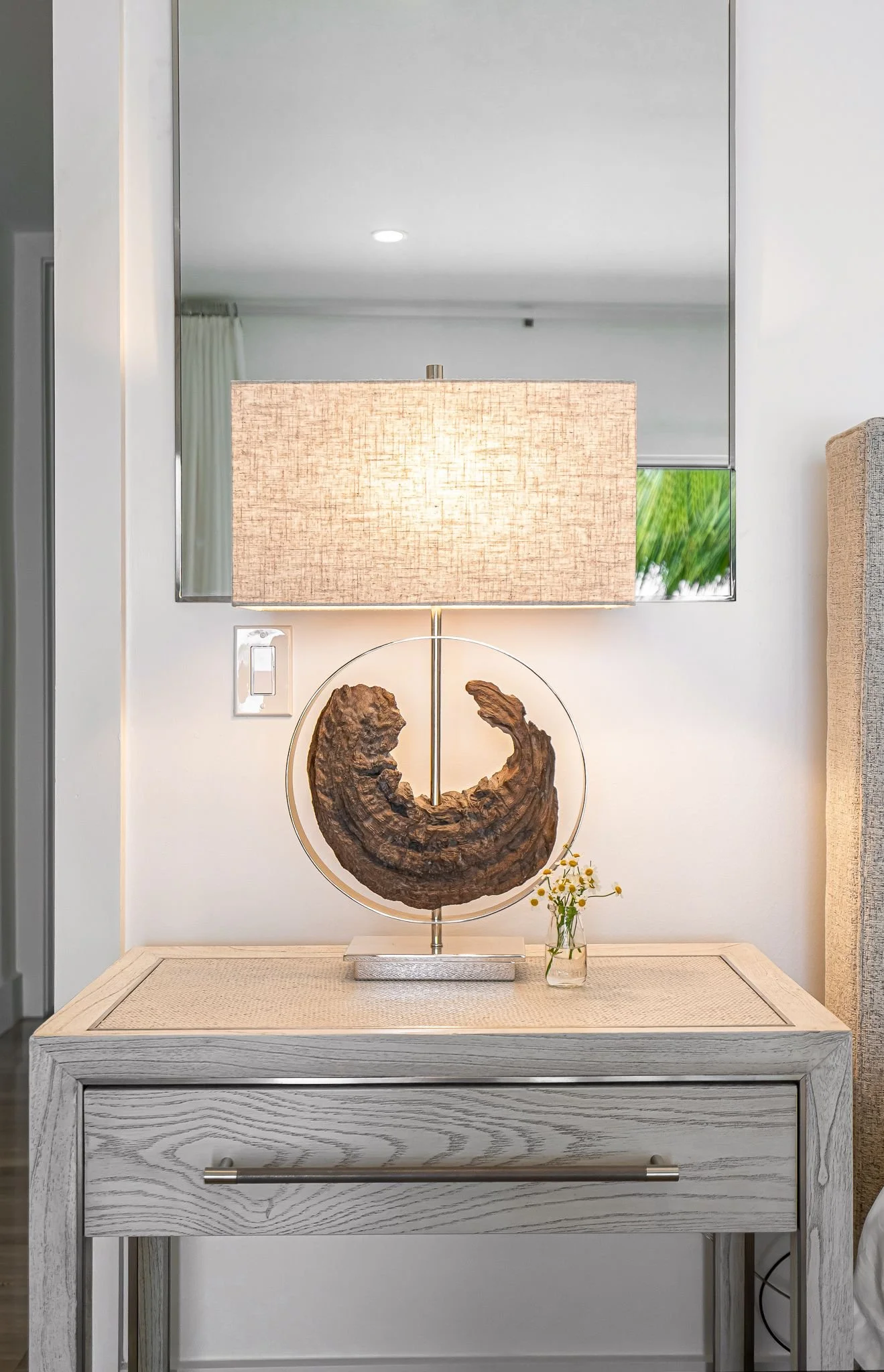 A bedside table with a beige woven top, decorated with a lamp featuring a circular wooden piece and a fabric shade, and a small glass vase with yellow flowers.