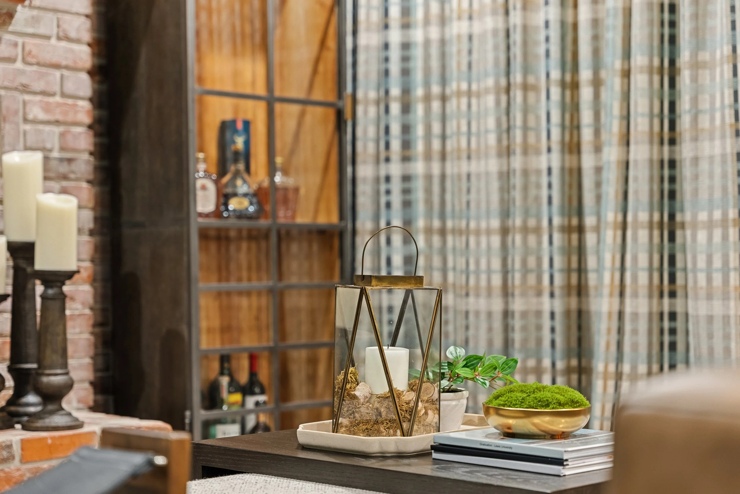 Decorative table centerpiece with a white candle inside a glass lantern, green plant, moss, and dried flowers, placed on a tray with magazines. Background includes a brick wall, candles, and a wooden cabinet with bottles, and patterned curtains.