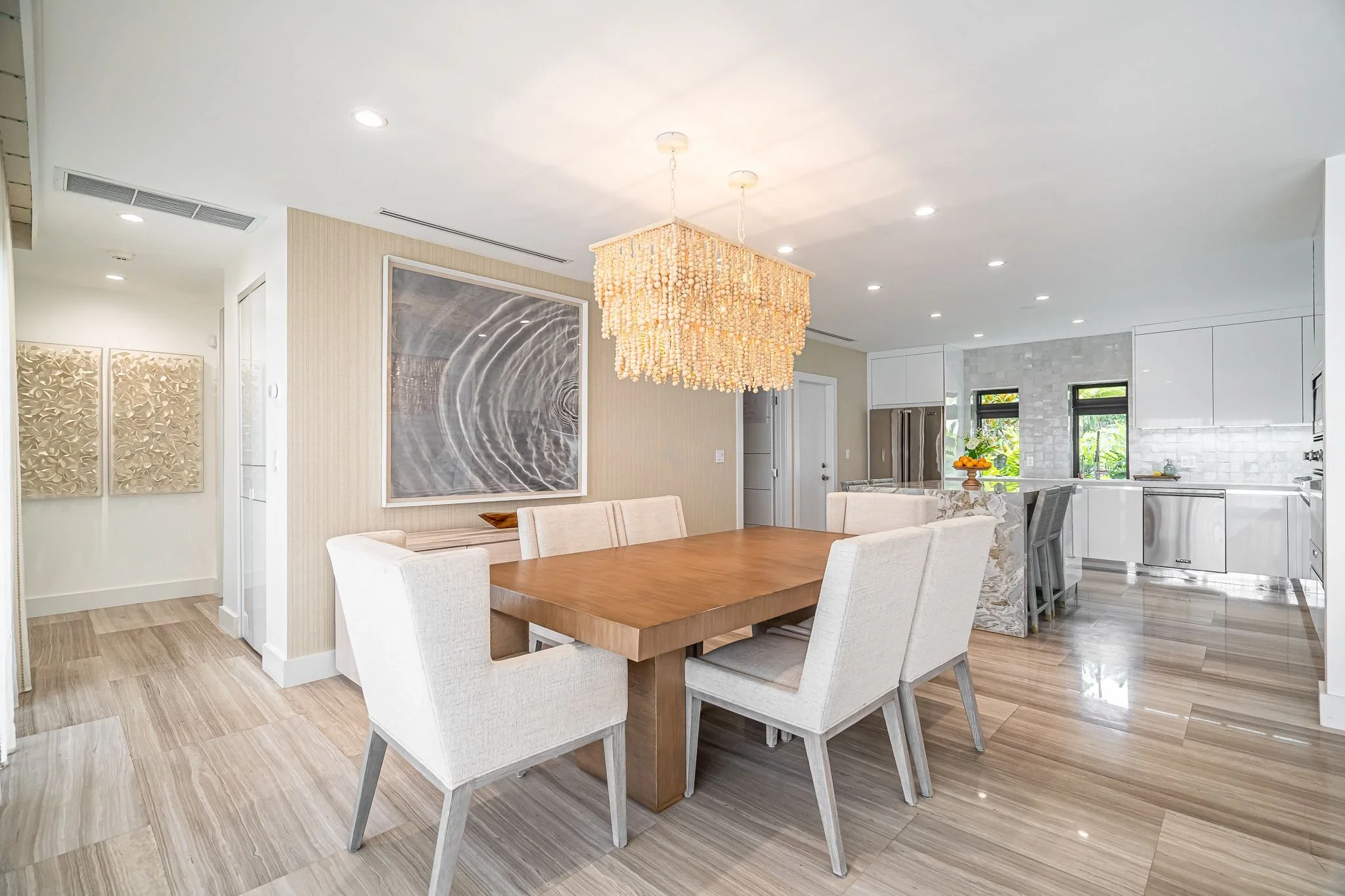 Modern open-concept dining and kitchen area with wooden dining table, upholstered chairs, and white cabinetry, illuminated by a hanging shell chandelier.