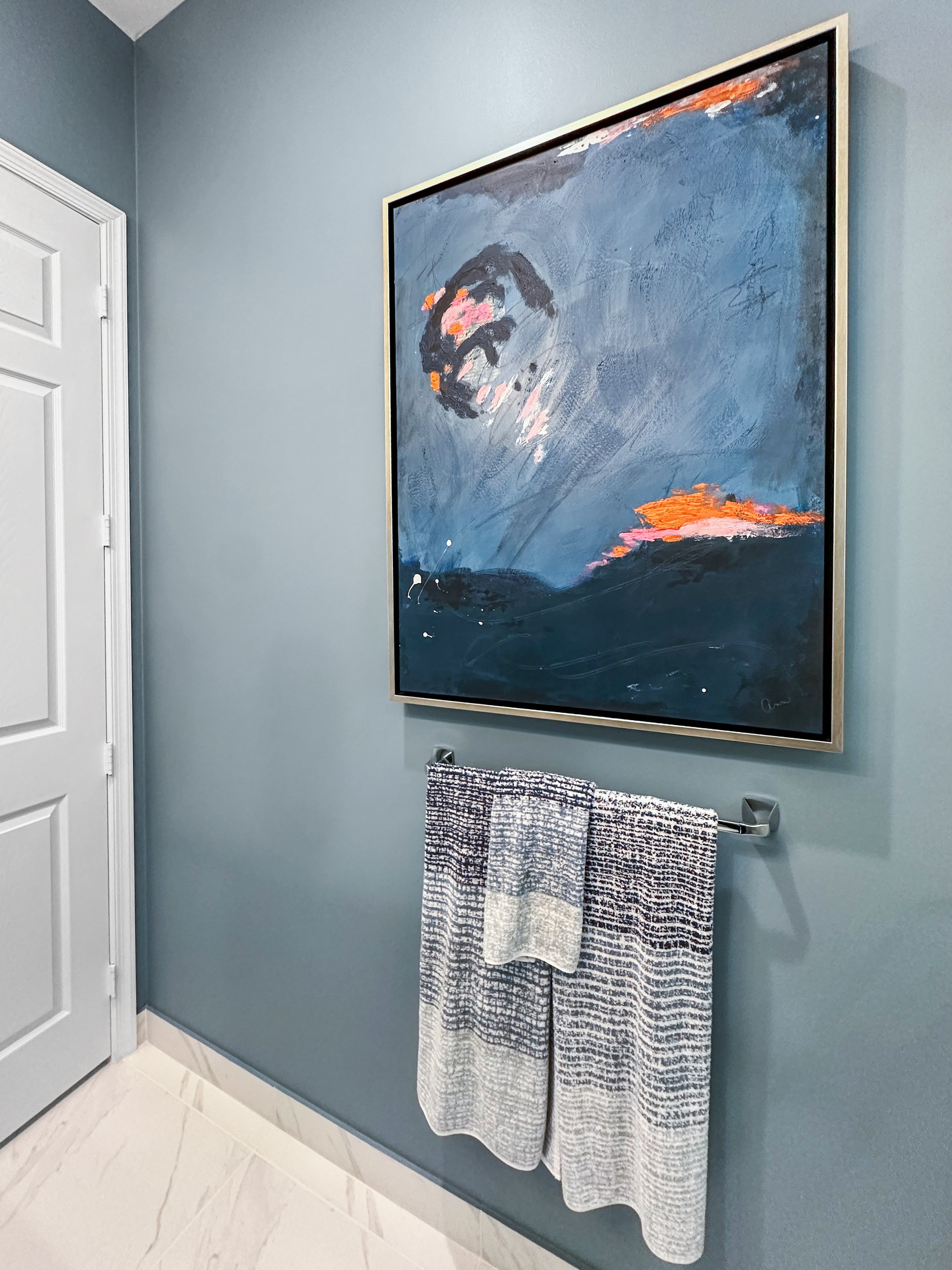 An abstract painting hangs on a gray wall above a towel bar with two patterned towels in a bathroom.