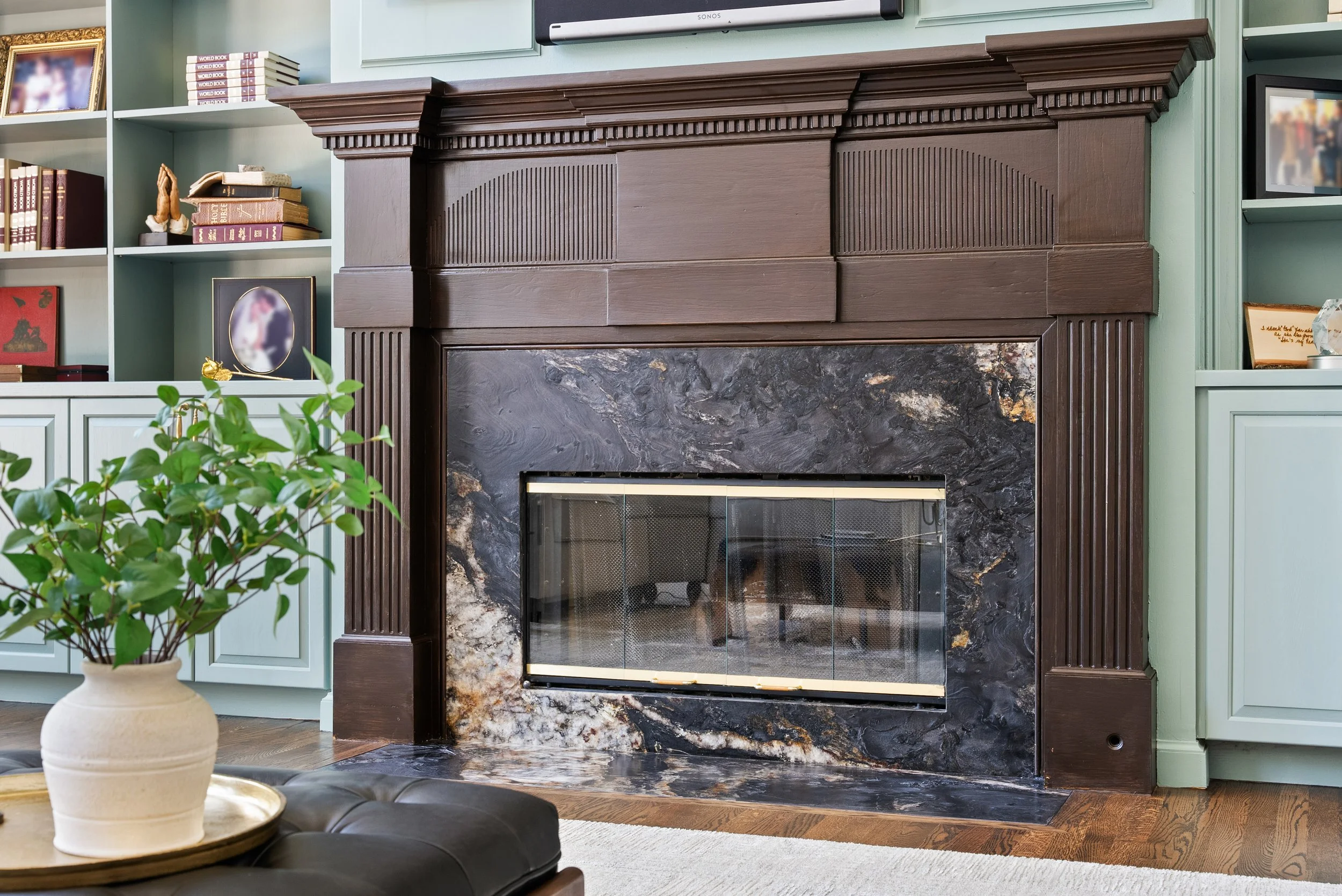 Living room fireplace with dark wood mantel and black marble surround, green built-in bookshelves on either side filled with books and decorative items, and a potted green plant on a wooden coffee table in the foreground.