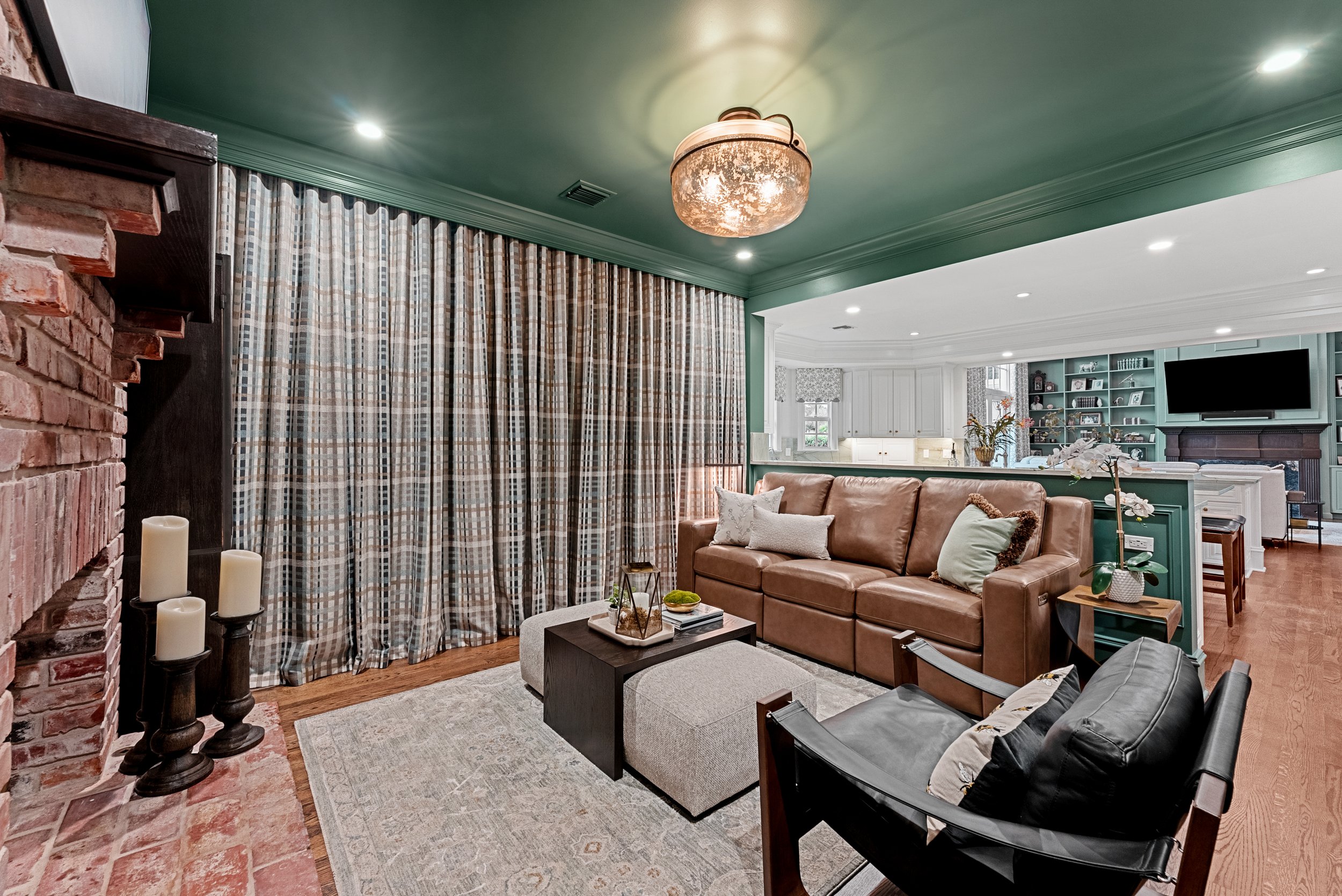 Spacious living room with green ceiling, brick fireplace, plaid curtains, brown leather sofa, black leather chair, light-colored ottomans, and houseplants, opening to a kitchen and dining area with built-in bookshelves and a television.