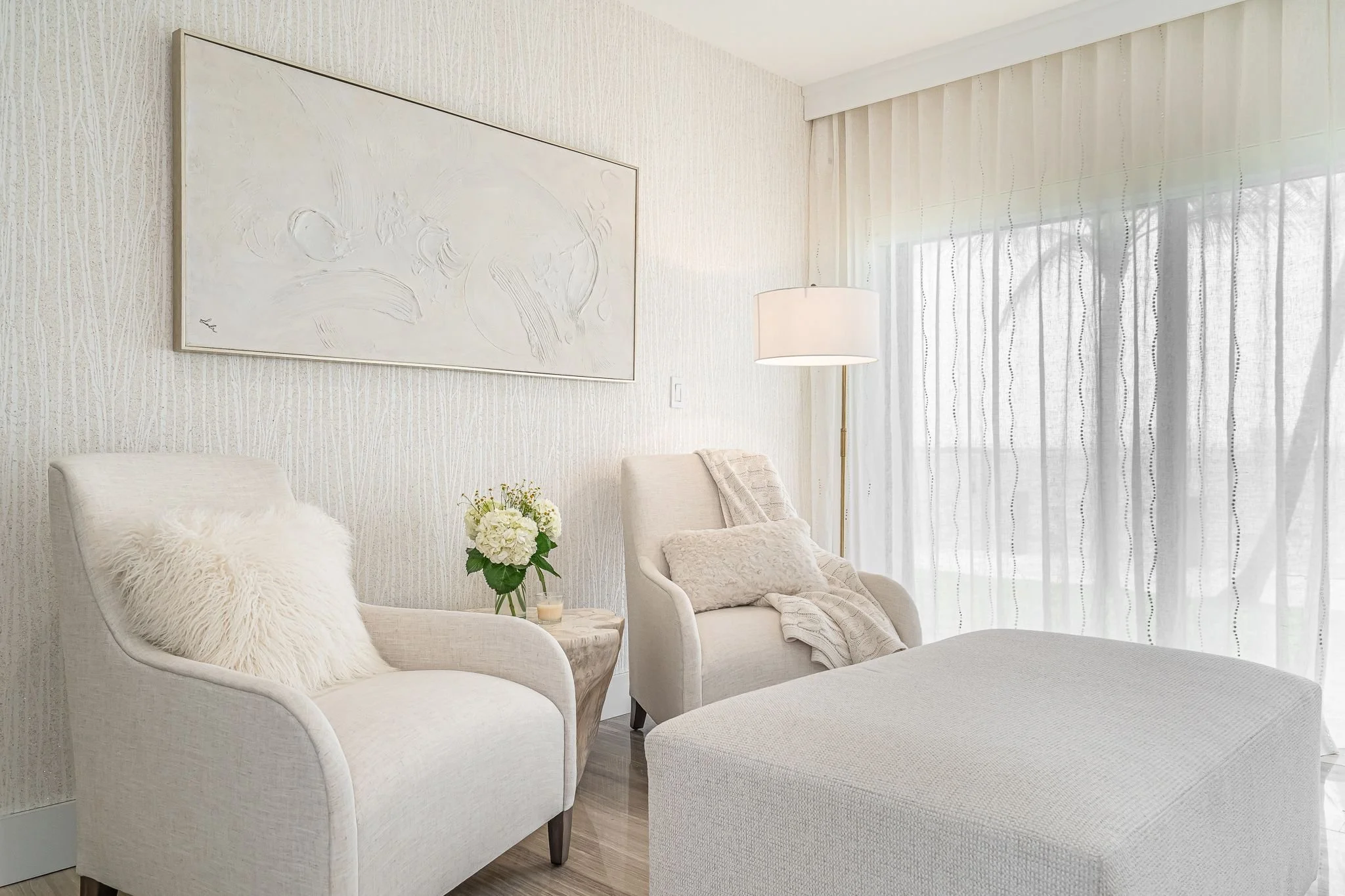 Bright living room with white upholstered armchairs, a side table with flowers, a floor lamp, and sheer curtains over large windows.