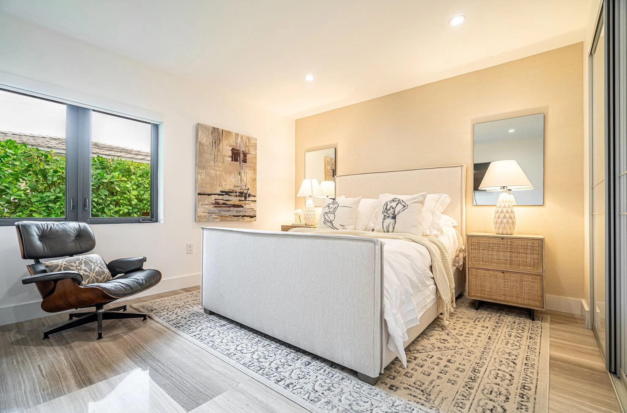 Bedroom with a cream upholstered bed, bedside tables with lamps, abstract wall art, a black leather lounge chair, and a window with greenery outside.