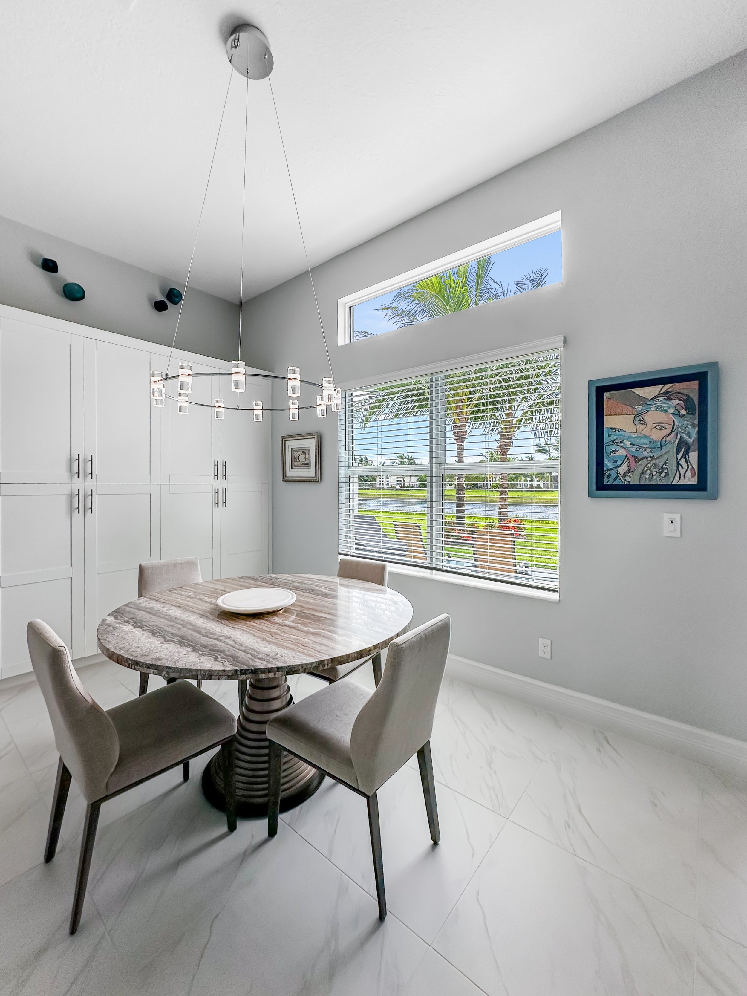 Dining room with round marble table, four beige chairs, white cabinetry, large window with blinds, palm trees outside, modern chandelier, and framed artwork on the wall.