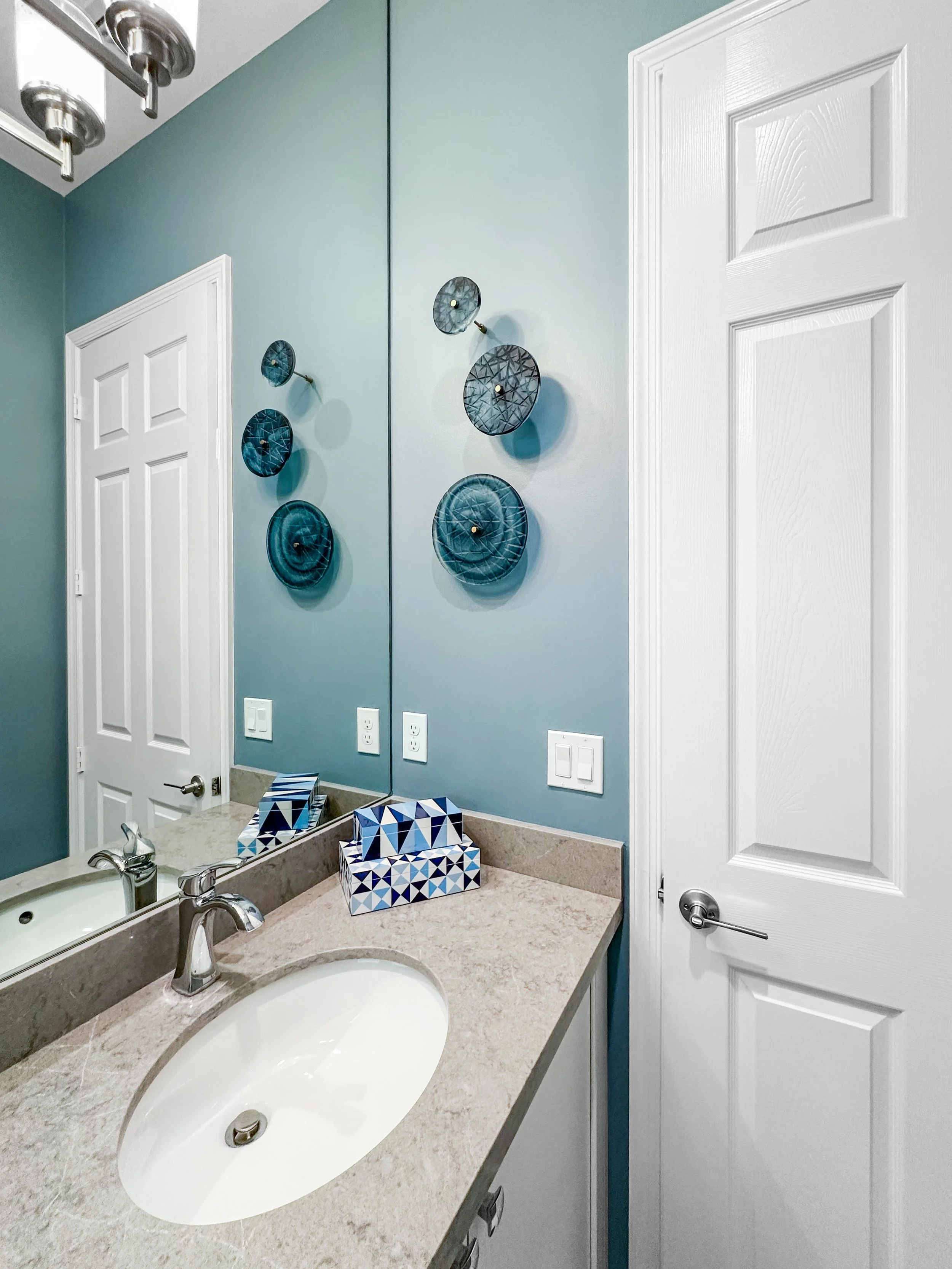 Bathroom with a beige countertop, white sink, and silver faucet, decorated with a blue and white tissue box with geometric patterns. Blue wall art made of circular disks in various shades of blue hung above the countertop. White door with a silver ha