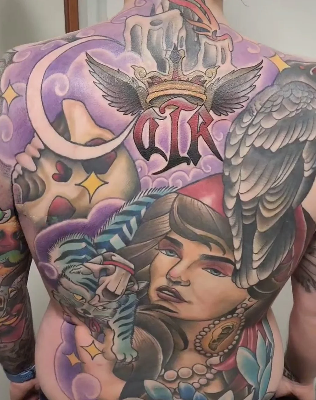 Colorful tattoo covering the person's back featuring a woman's face, a tiger, a snake, a crown, clouds, and various decorative elements.