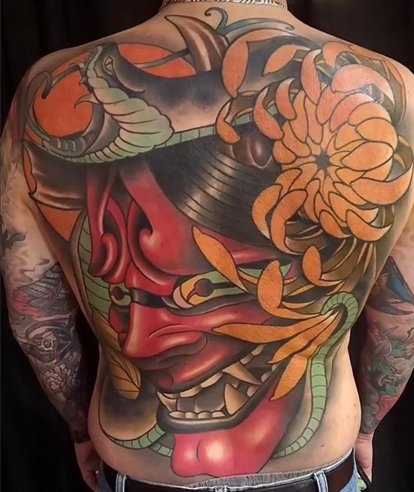 Colorful tattoo covering the back of a person, featuring a traditional Japanese Hannya mask with a red face, sharp teeth, and horns, surrounded by orange and green elements, including a dragon, cloud-like patterns, and floral motifs.
