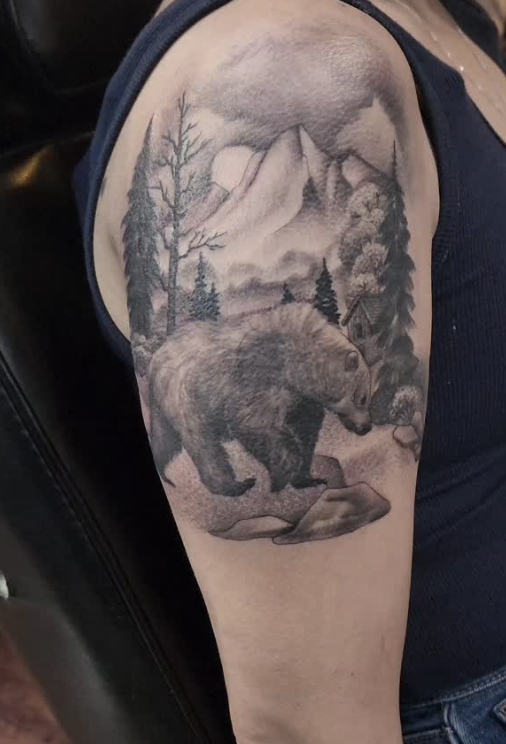 Tattoo of a bear walking among rocks with mountains, pine trees, and a cabin in the background on a person's upper arm.