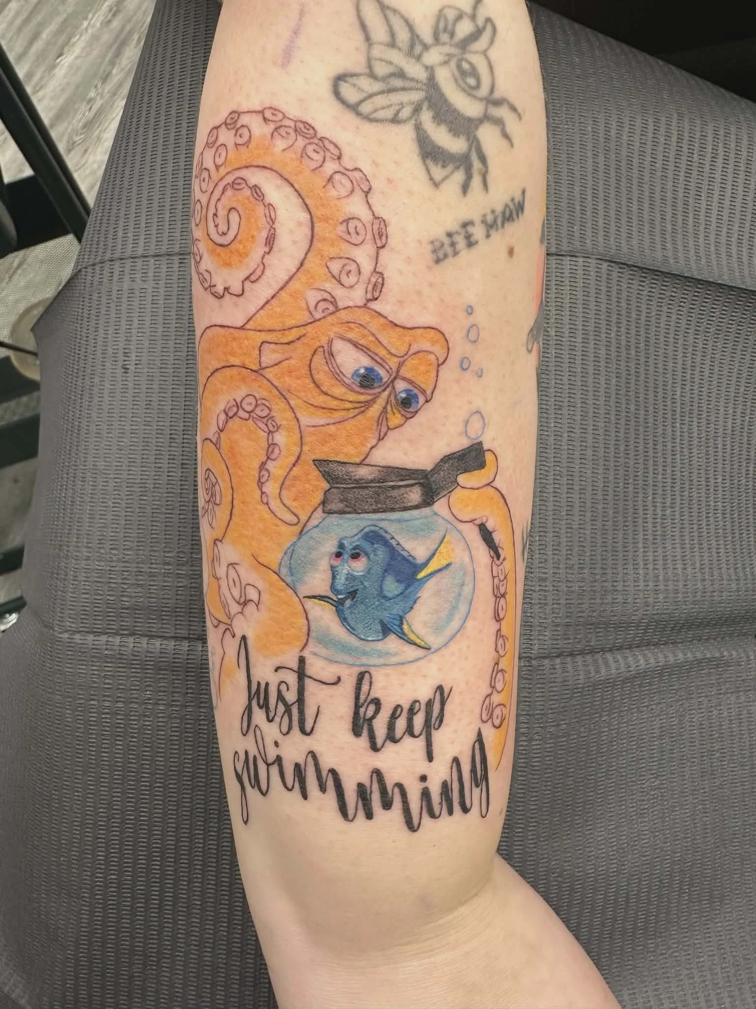 A colorful tattoo on a person's arm featuring a cartoon octopus with glasses, a fish in a fishbowl, and a bee at the top. The tattoo includes the phrase "Just keep swimming" in cursive text.