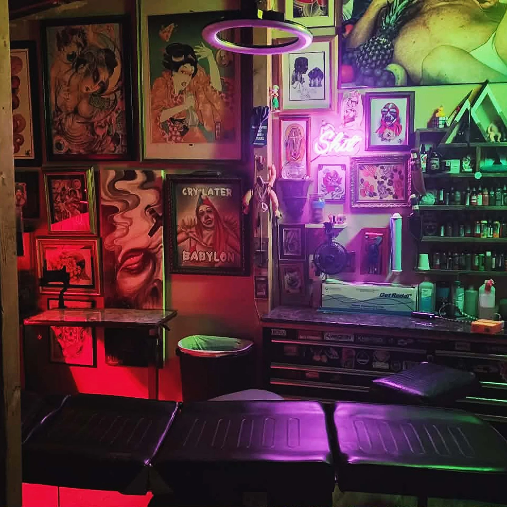 A dimly lit room decorated with vibrant, colorful artwork and neon signs. The walls are adorned with various framed pictures, and there is a neon "Sake" sign glowing pink. There is a small sink with shelves of bottles and containers nearby. The seating area includes a black leather bench in the foreground, with a small table and a trash can nearby.